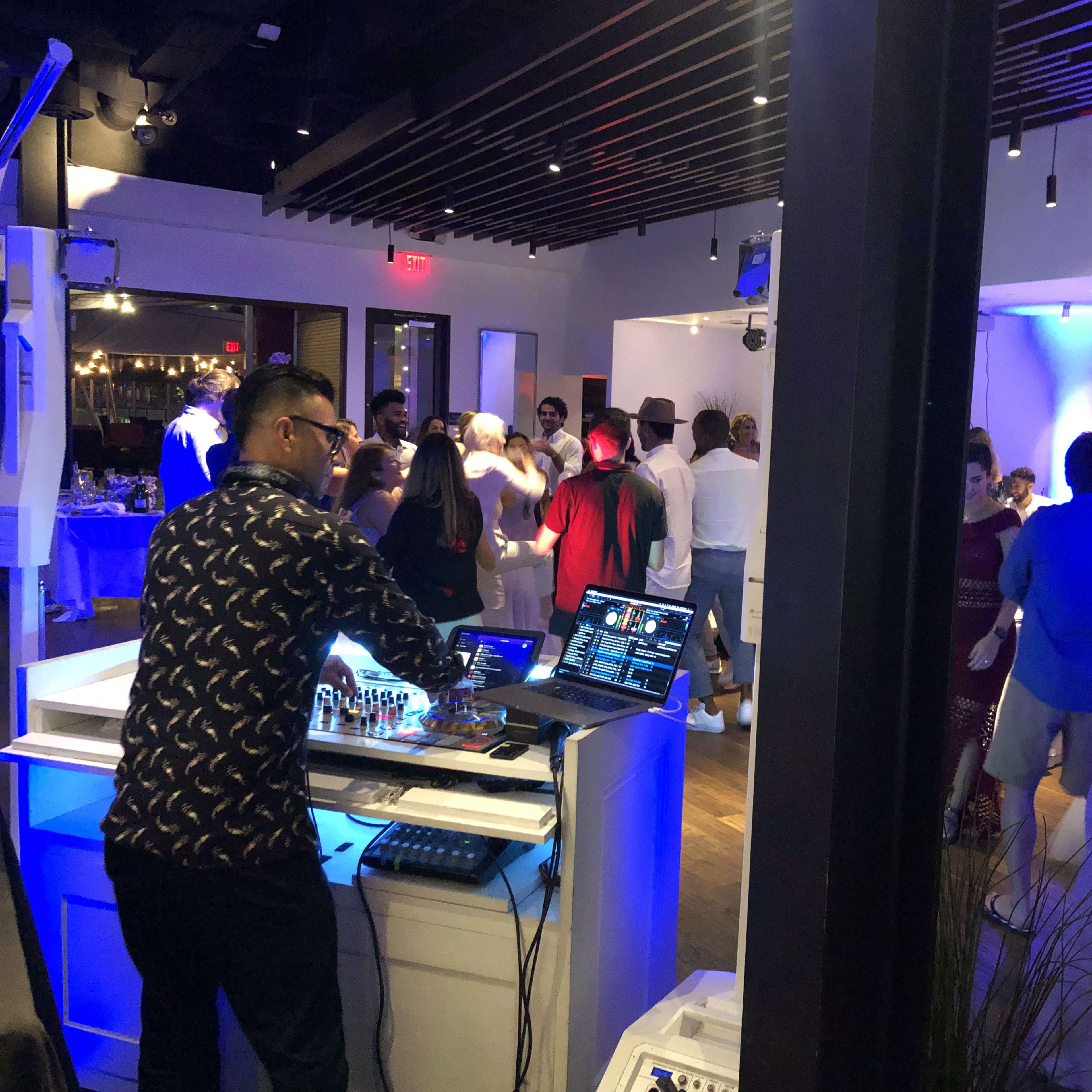 A DJ mixes music at a lively dance party in a modern venue filled with guests dancing and socializing.
