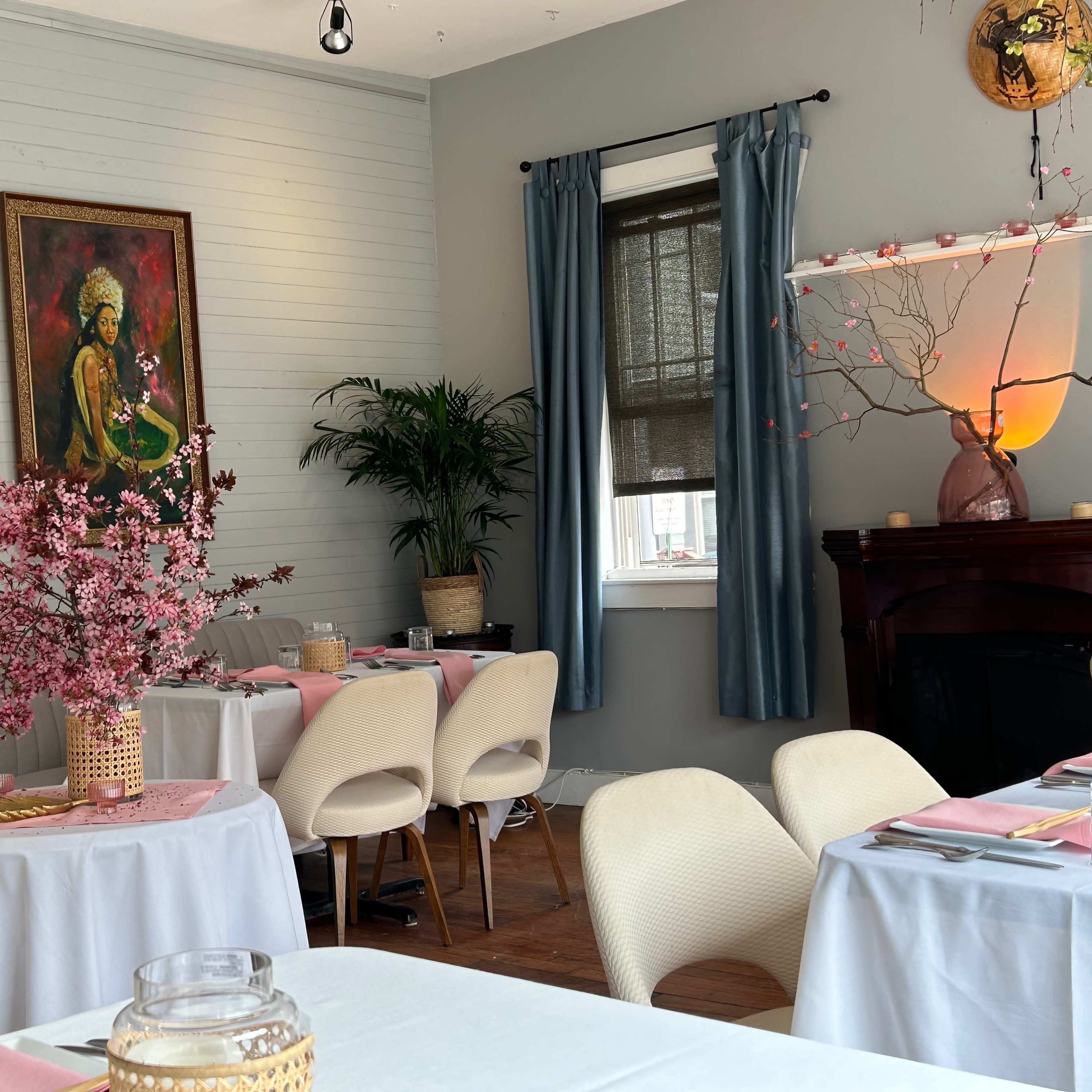 A dining area with white tables, a potted plant, and a large piece of artwork on the wall, complemented by floral centerpieces and light blue curtains.