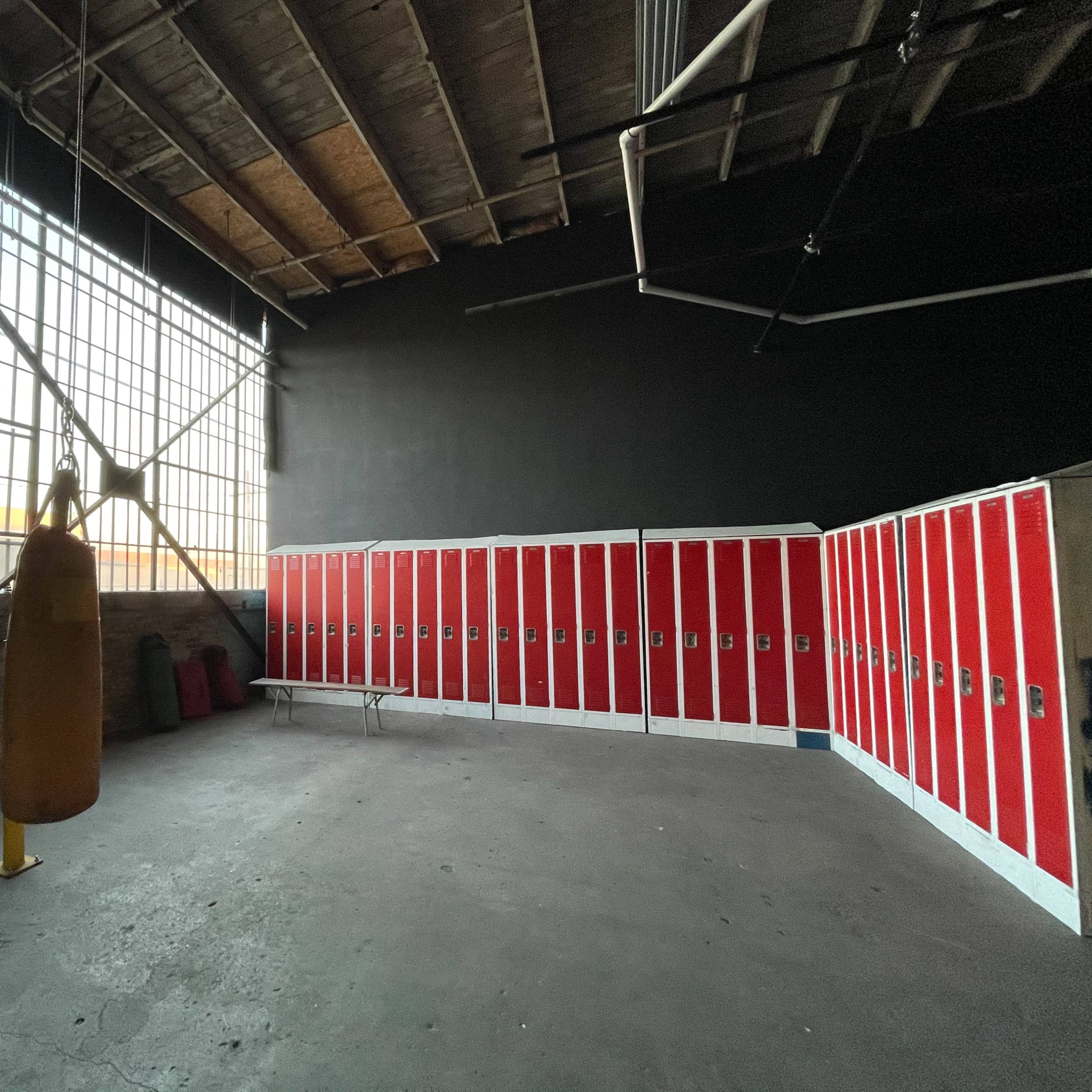 A sparsely decorated gym interior featuring a heavy punching bag suspended from the ceiling and a row of red lockers along one wall.