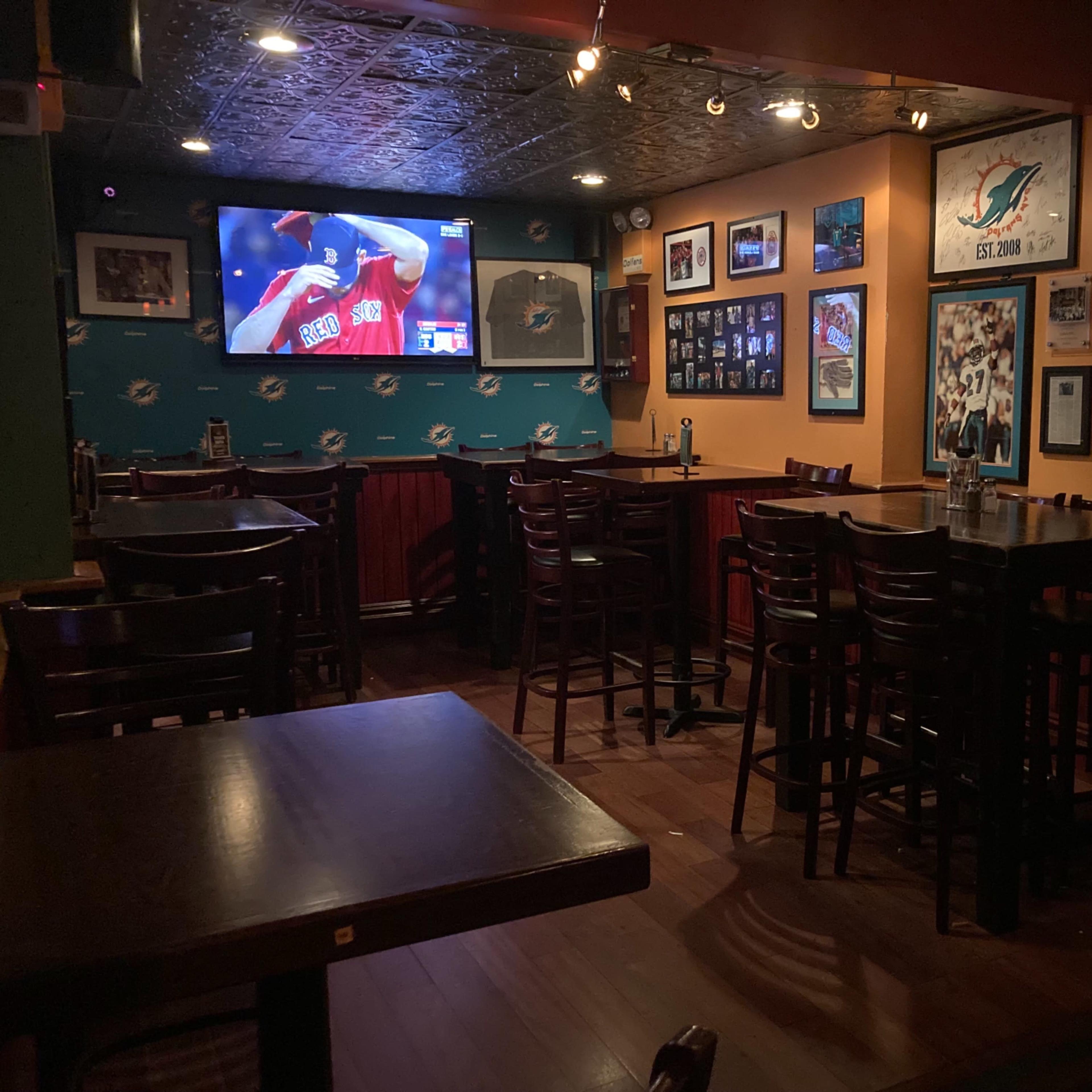 The interior of a bar with wooden tables and chairs, featuring a large screen displaying a sports game and walls adorned with framed photographs.