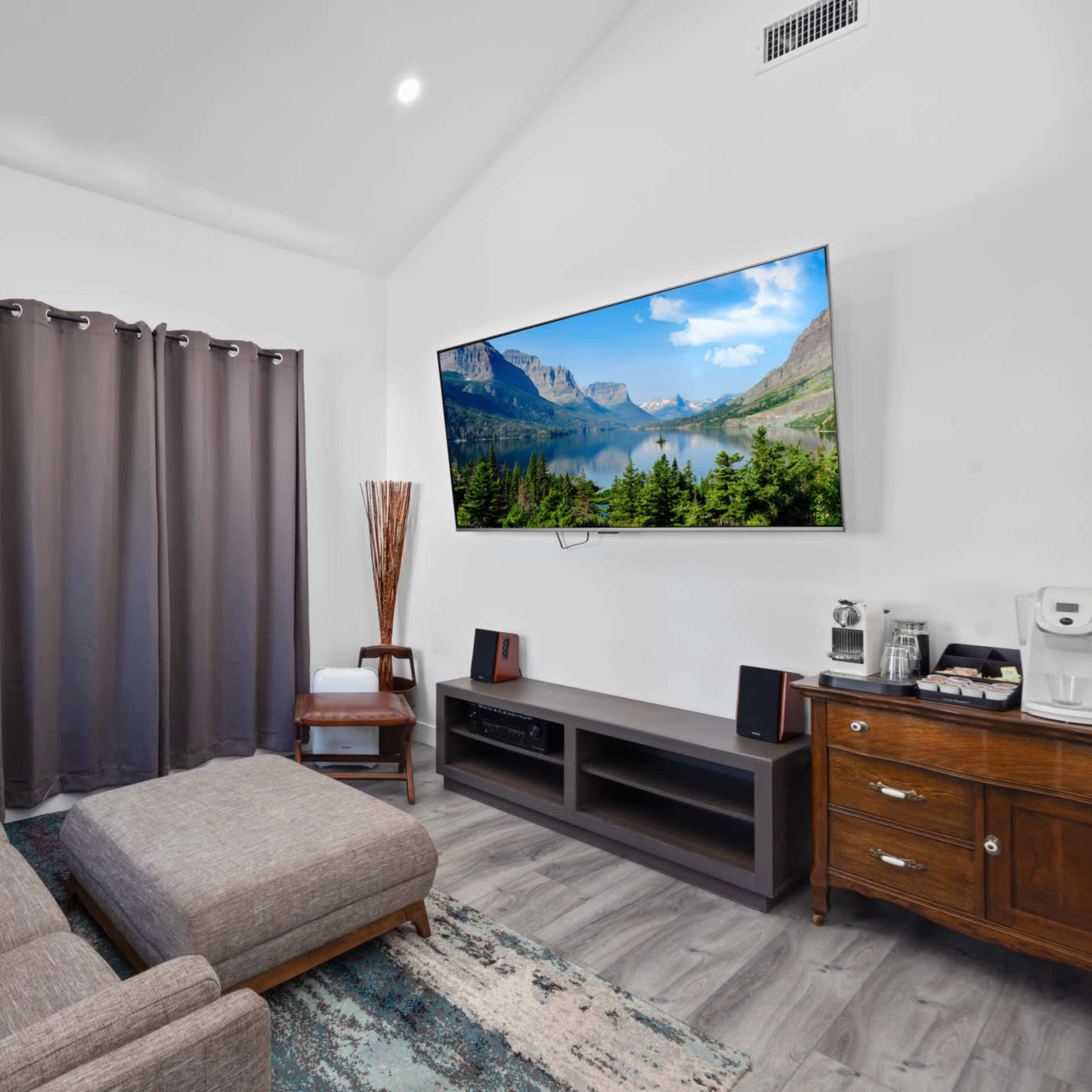 The living room features a gray sectional sofa, a large wall-mounted television displaying a scenic landscape, and dark curtains beside a wooden cabinet.