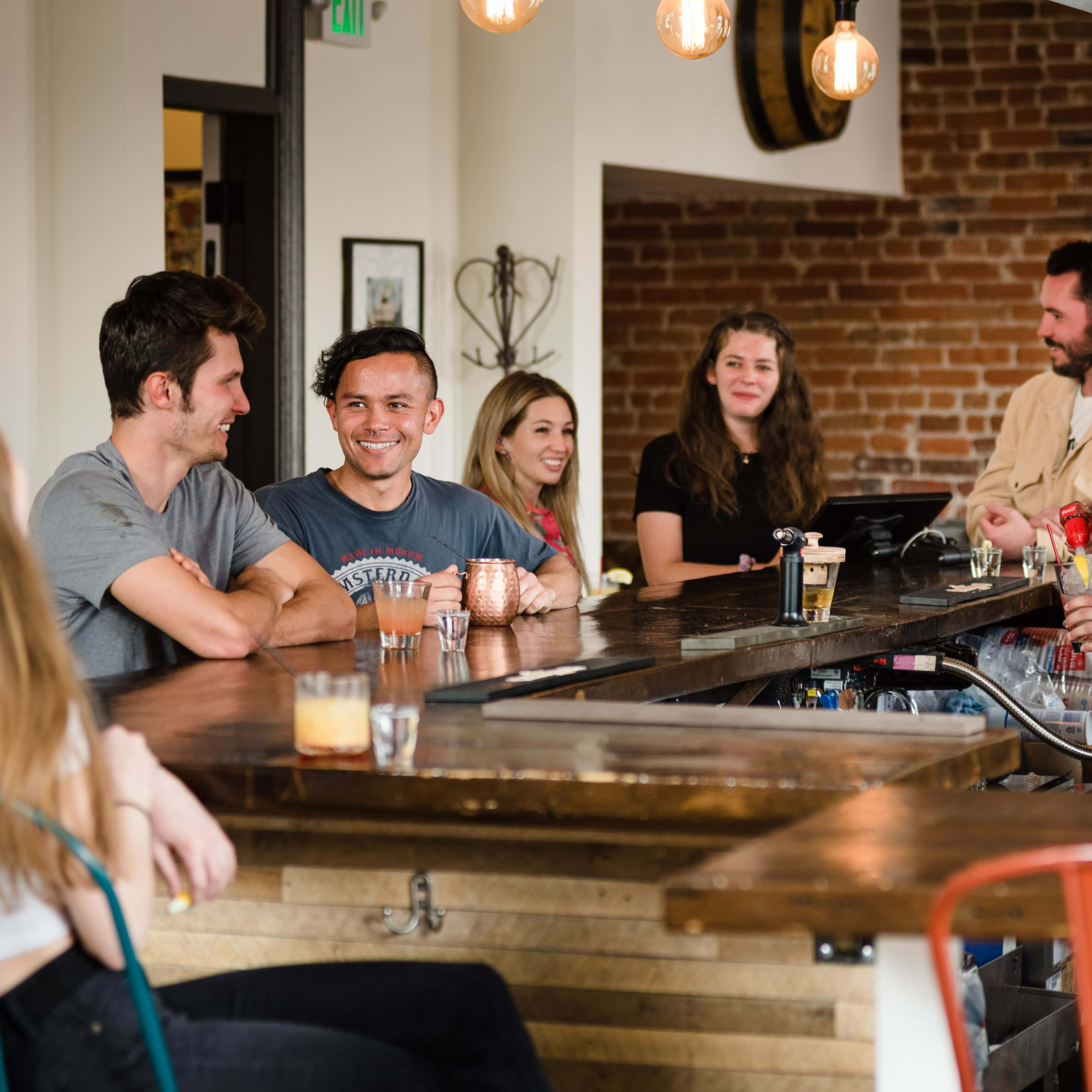 A group of people is gathered at a bar, interacting and enjoying drinks in a warmly lit space with exposed brick walls.