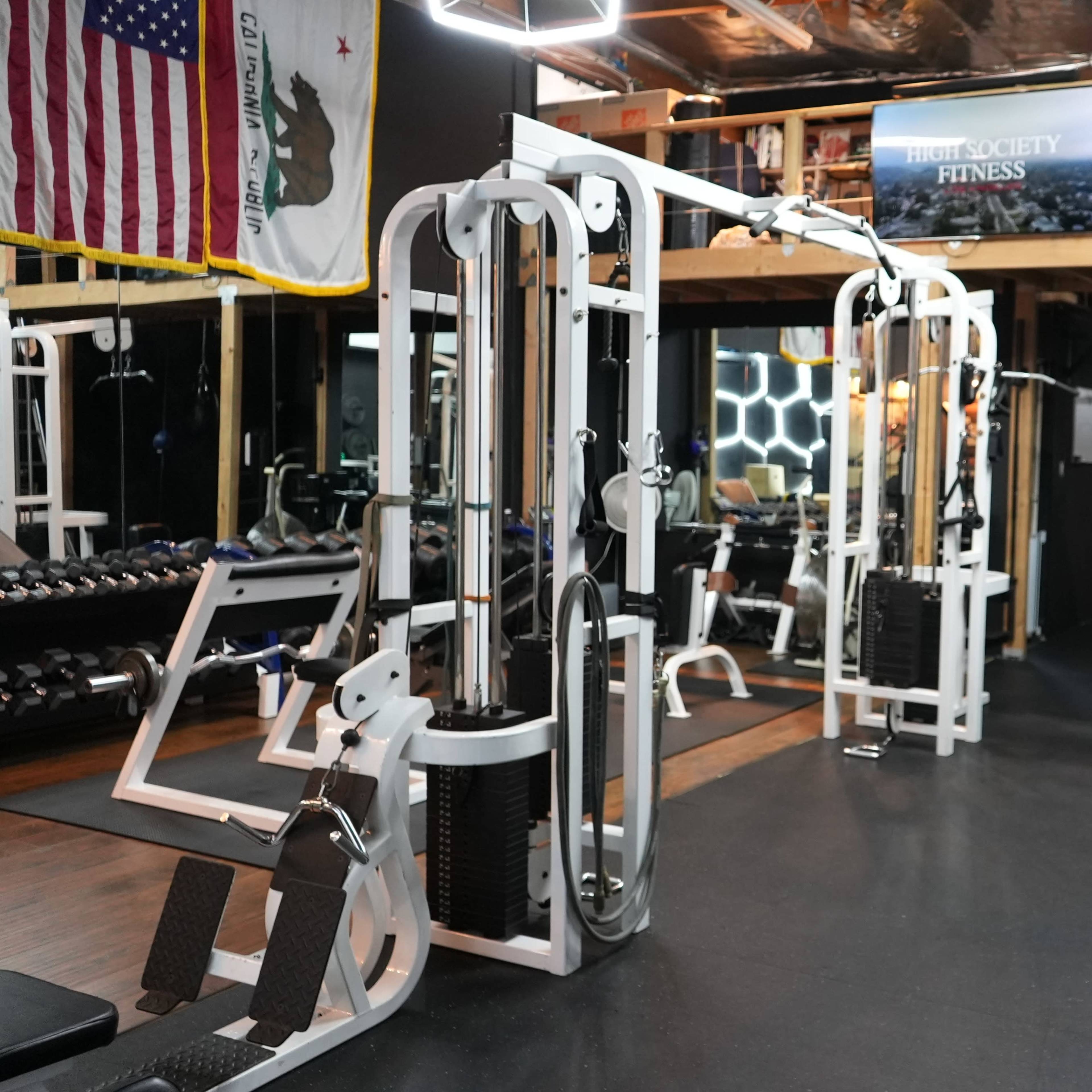 A well-equipped gym featuring various weight training machines, dumbbells, and a mirrored wall, with flags and a sign visible in the background.