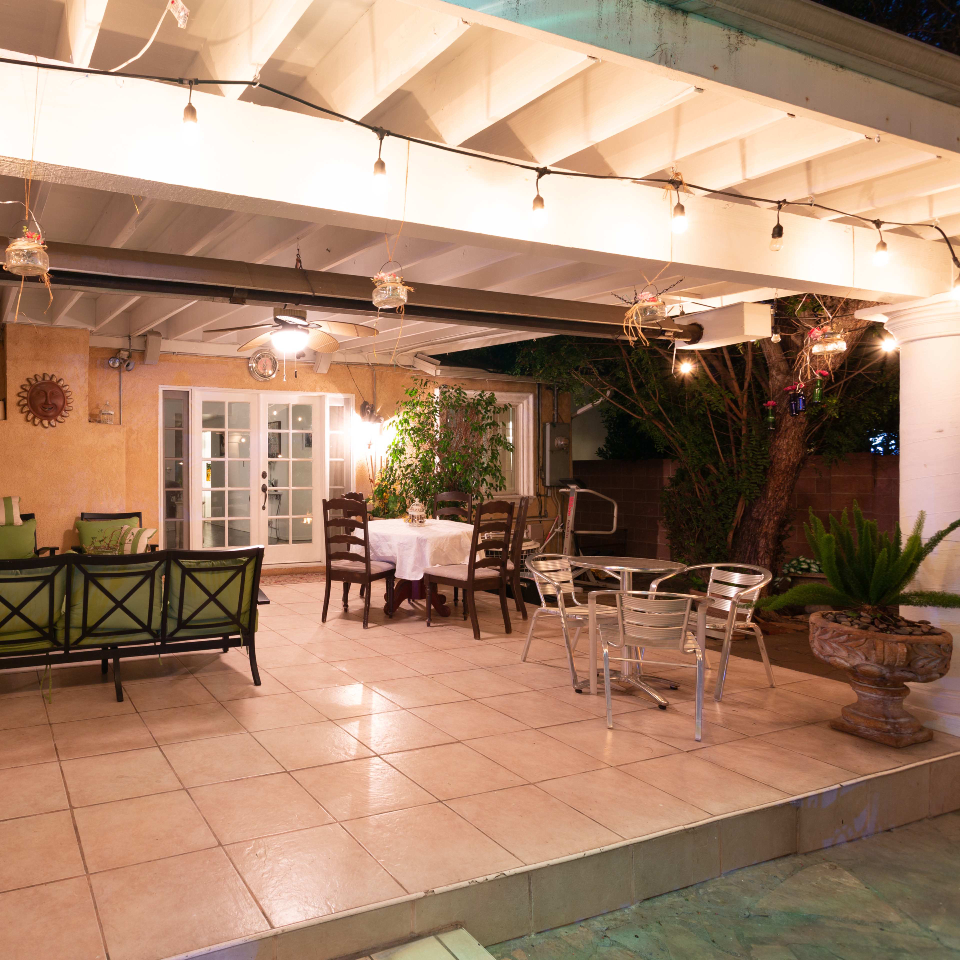 The scene shows a covered patio area with furniture, potted plants, and string lights.