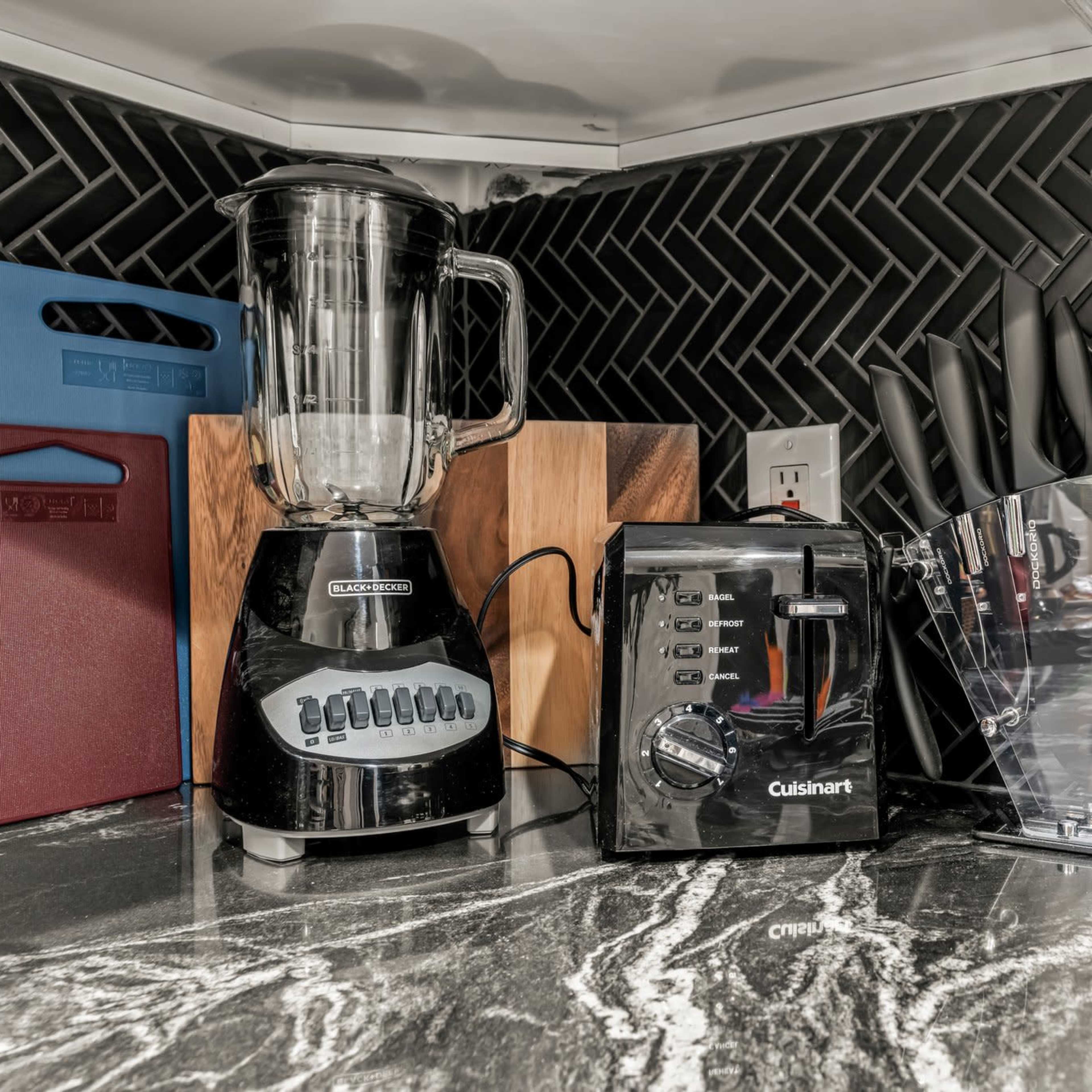 A kitchen countertop with a blender, a toaster, a knife block, and a cutting board against a herringbone-patterned backsplash.
