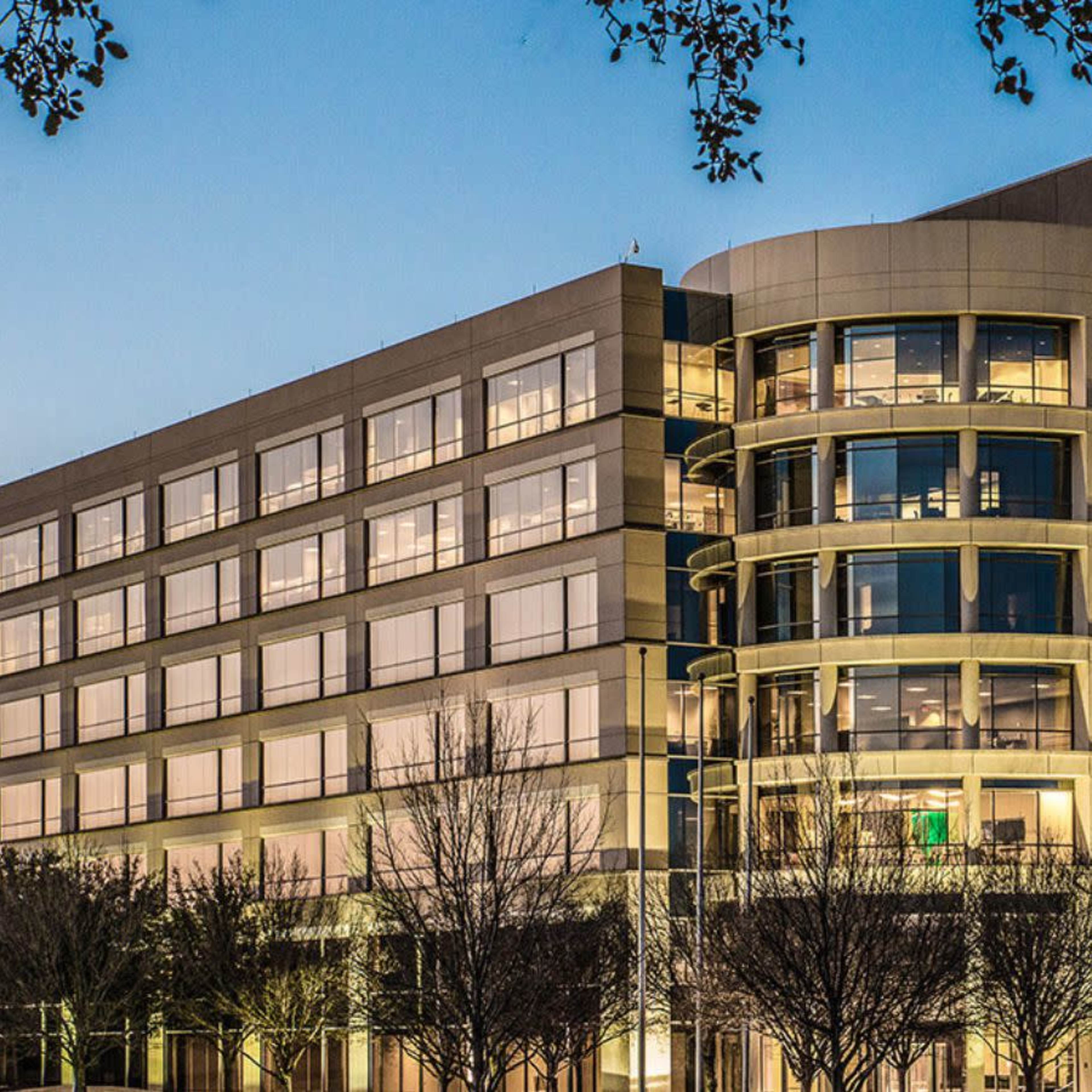 A modern, multi-story office building illuminated at dusk, surrounded by bare trees.
