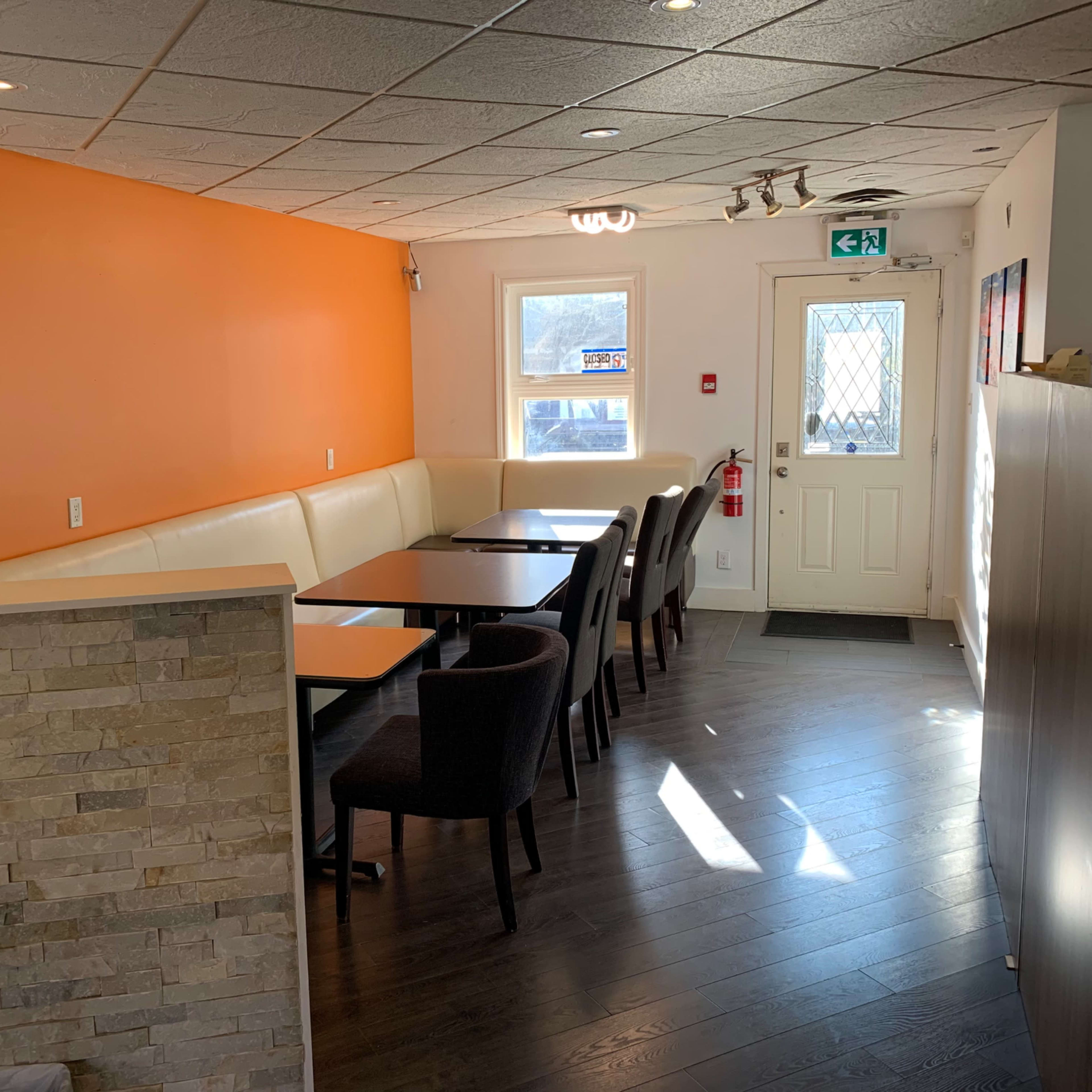 A small restaurant interior with a beige and orange color scheme, featuring a row of tables and chairs along one side and a large window near the entrance.