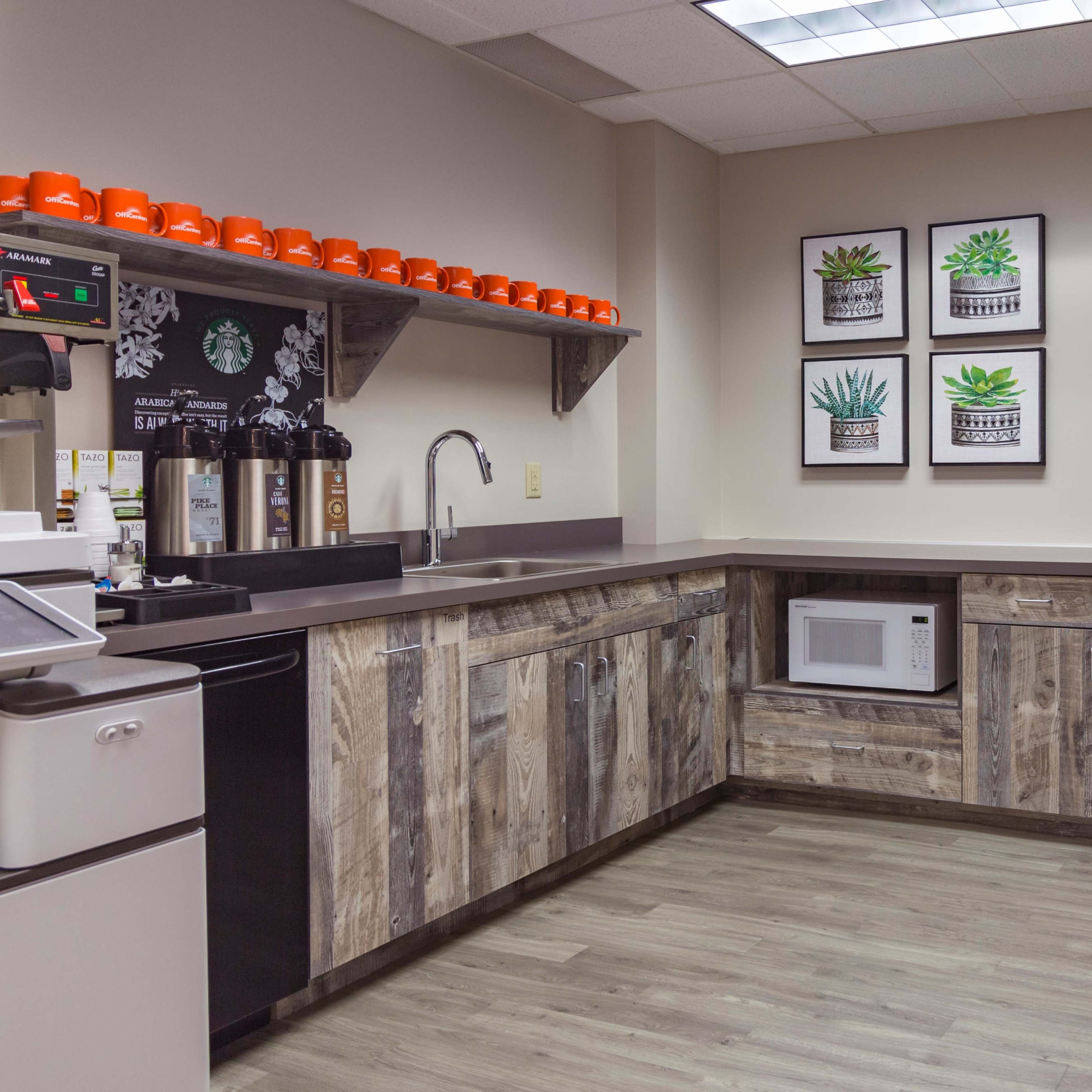A modern kitchen area featuring a refrigerator, microwave, and coffee station, with wooden cabinetry and framed plant artwork on the wall.