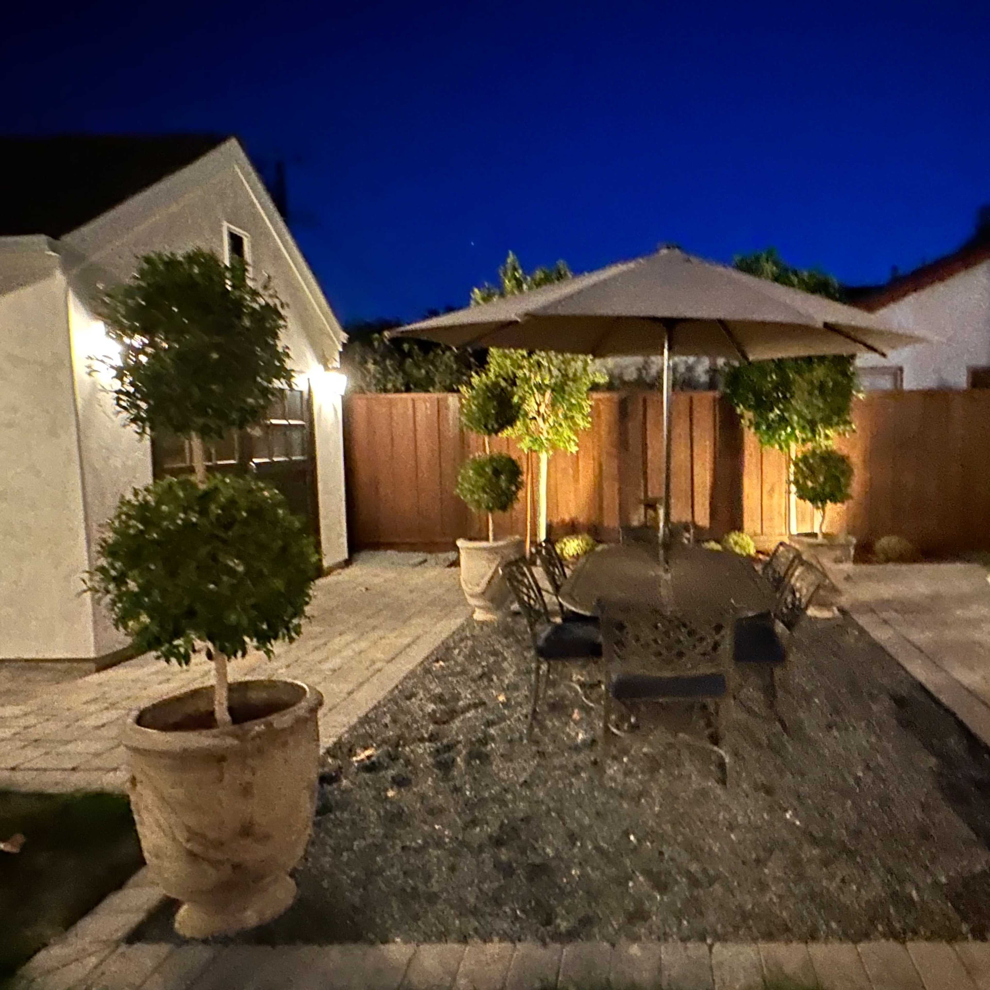 A patio features a table with chairs beneath a large umbrella, surrounded by potted trees and a gravel area in a backyard at night.