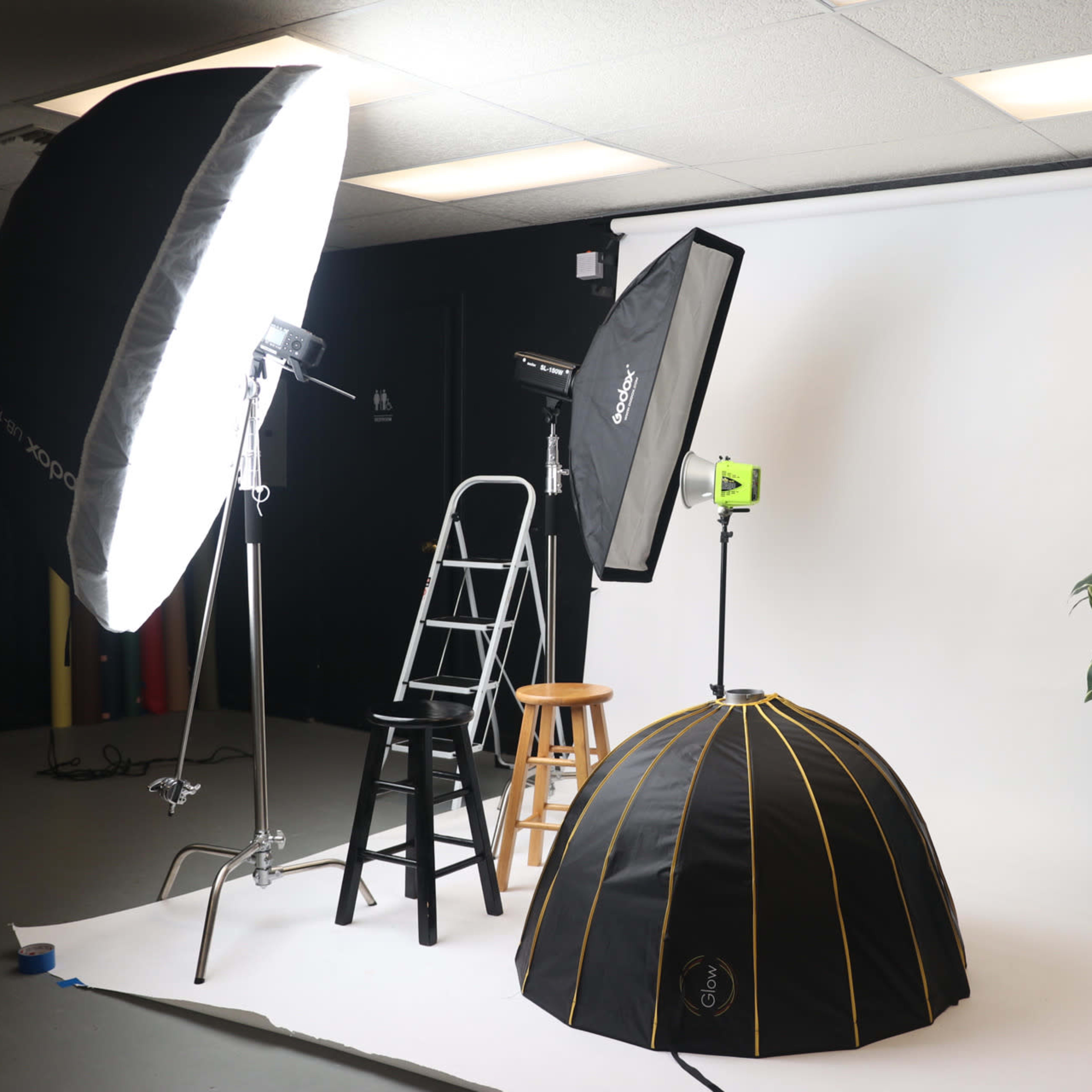 A photography studio setup featuring a white backdrop, various lighting equipment, a ladder, a black stool, a plant, and a light modifier.