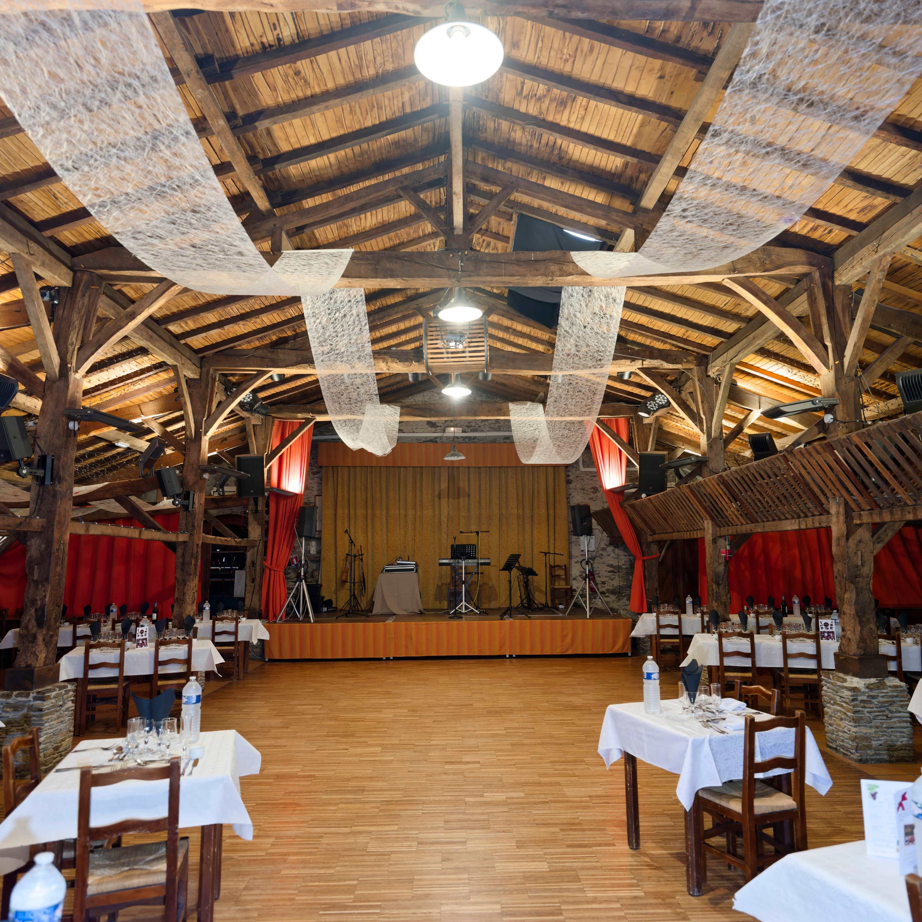 A spacious restaurant interior with wooden beams, tables set for dining, and a small stage at one end.