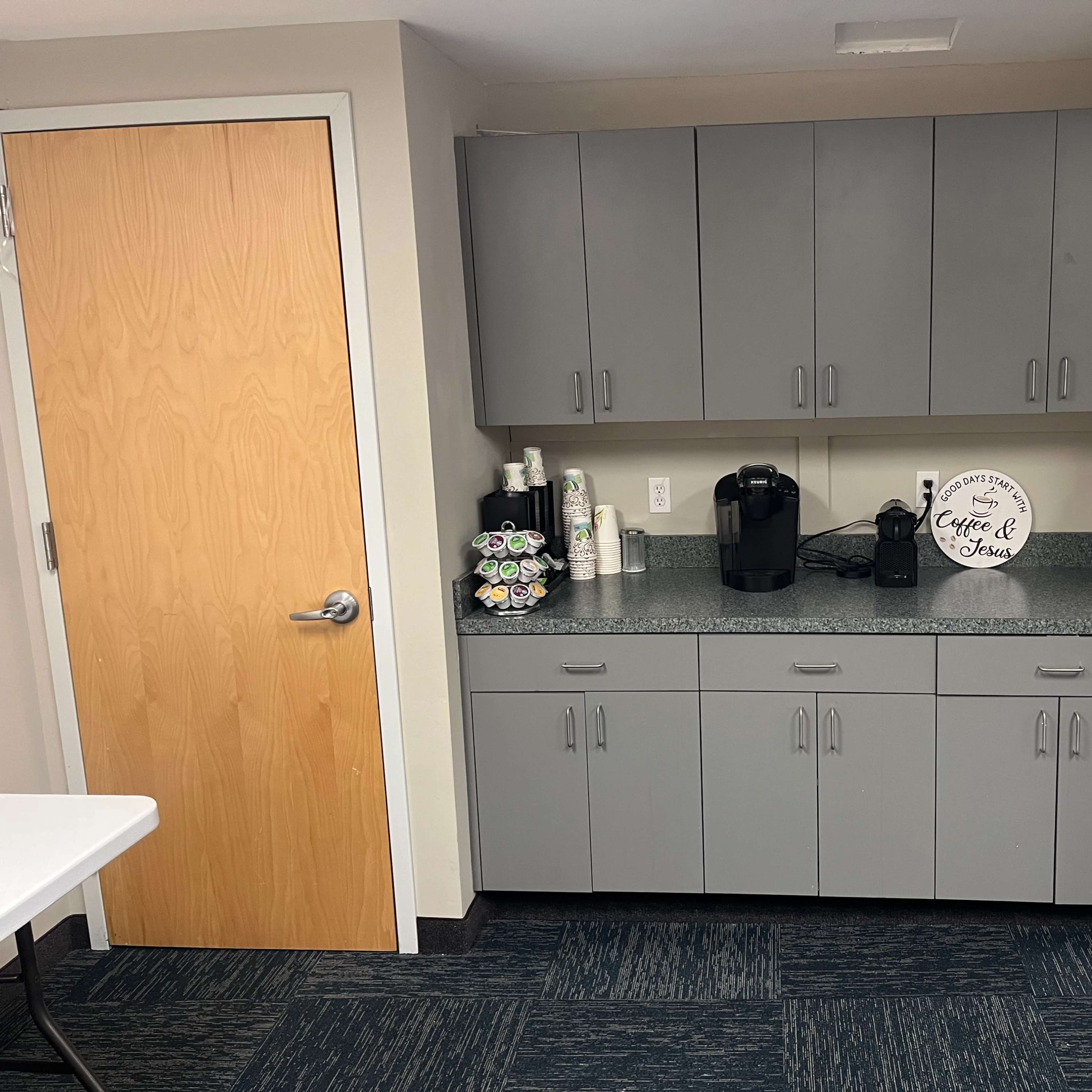 A gray cabinet area with a countertop, featuring a coffee maker, cups, and a decorative sign, located near two doors.