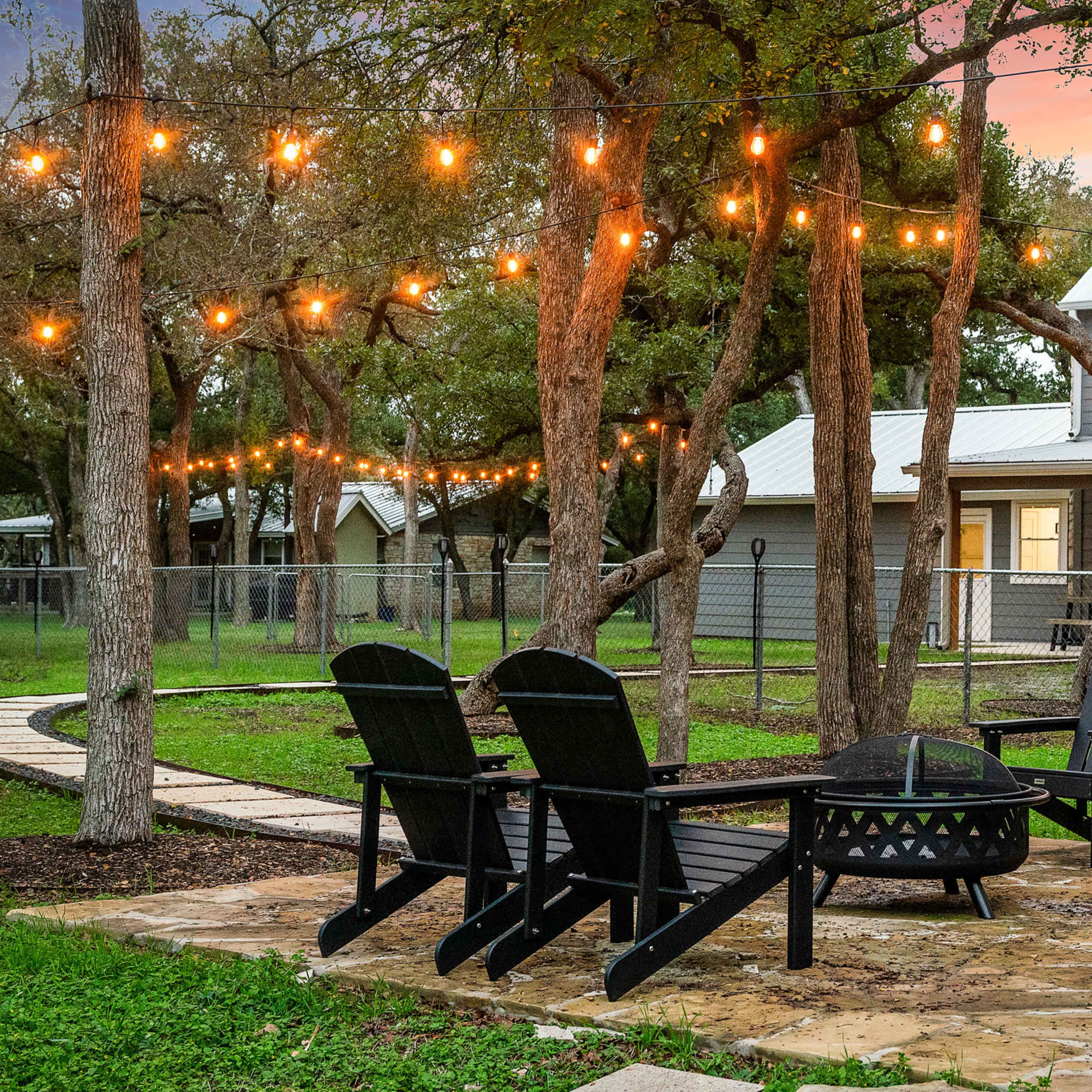 A circular stone patio with several black Adirondack chairs surrounds a fire pit, illuminated by string lights draped among the trees in a backyard setting.
