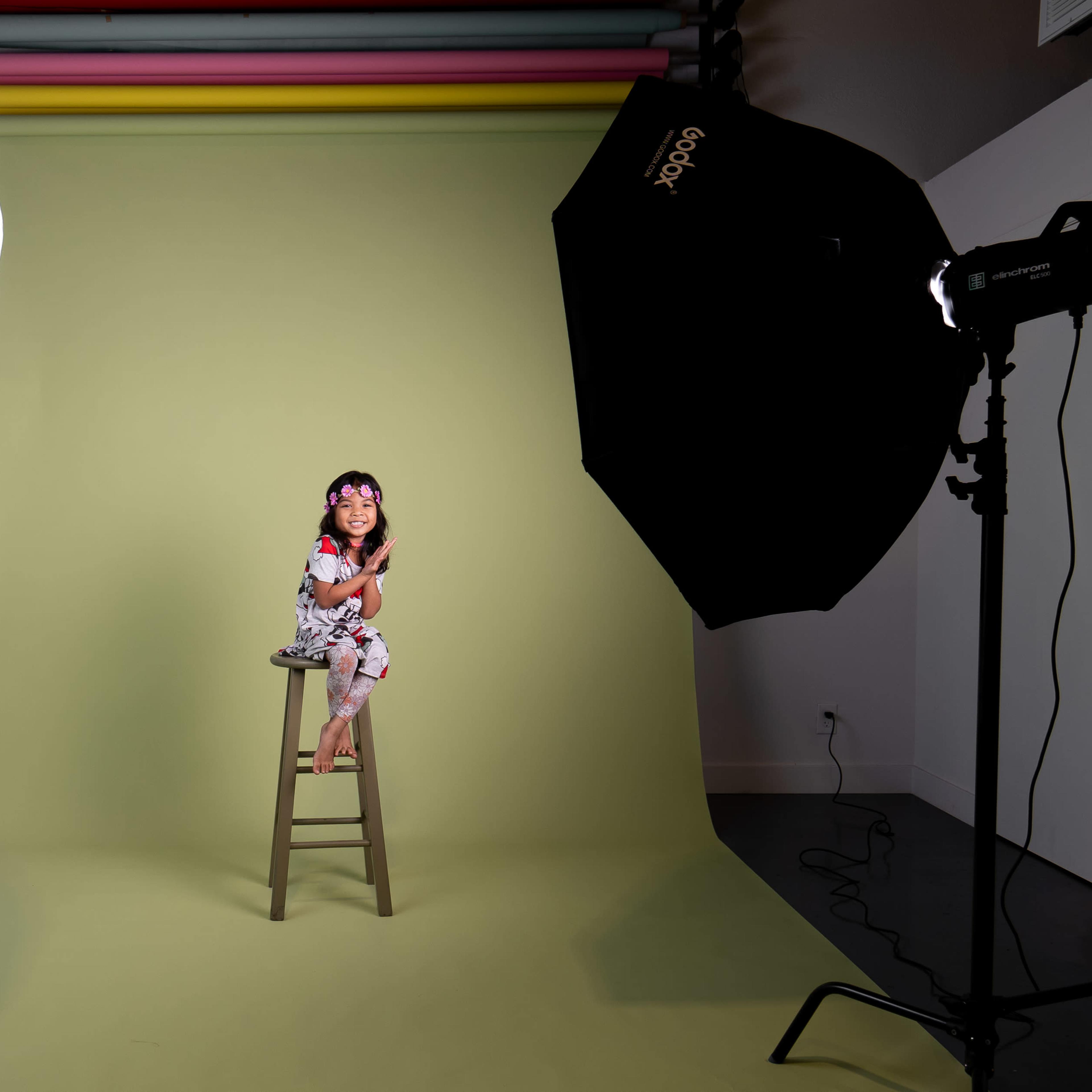 A child sits on a wooden stool in front of a green backdrop, illuminated by two large studio lights.