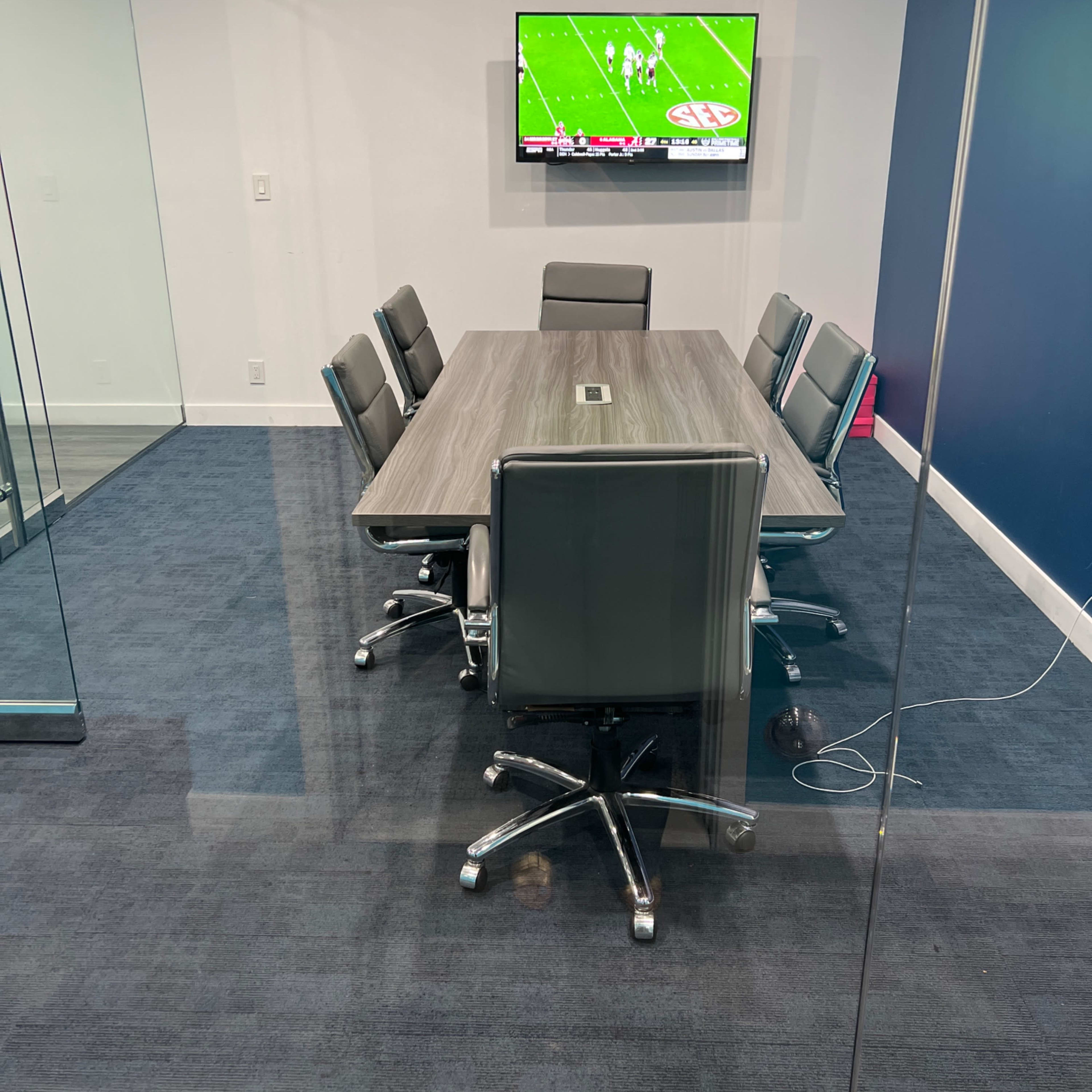A conference room with a large table surrounded by six chairs, featuring a television mounted on the wall displaying a sports game.
