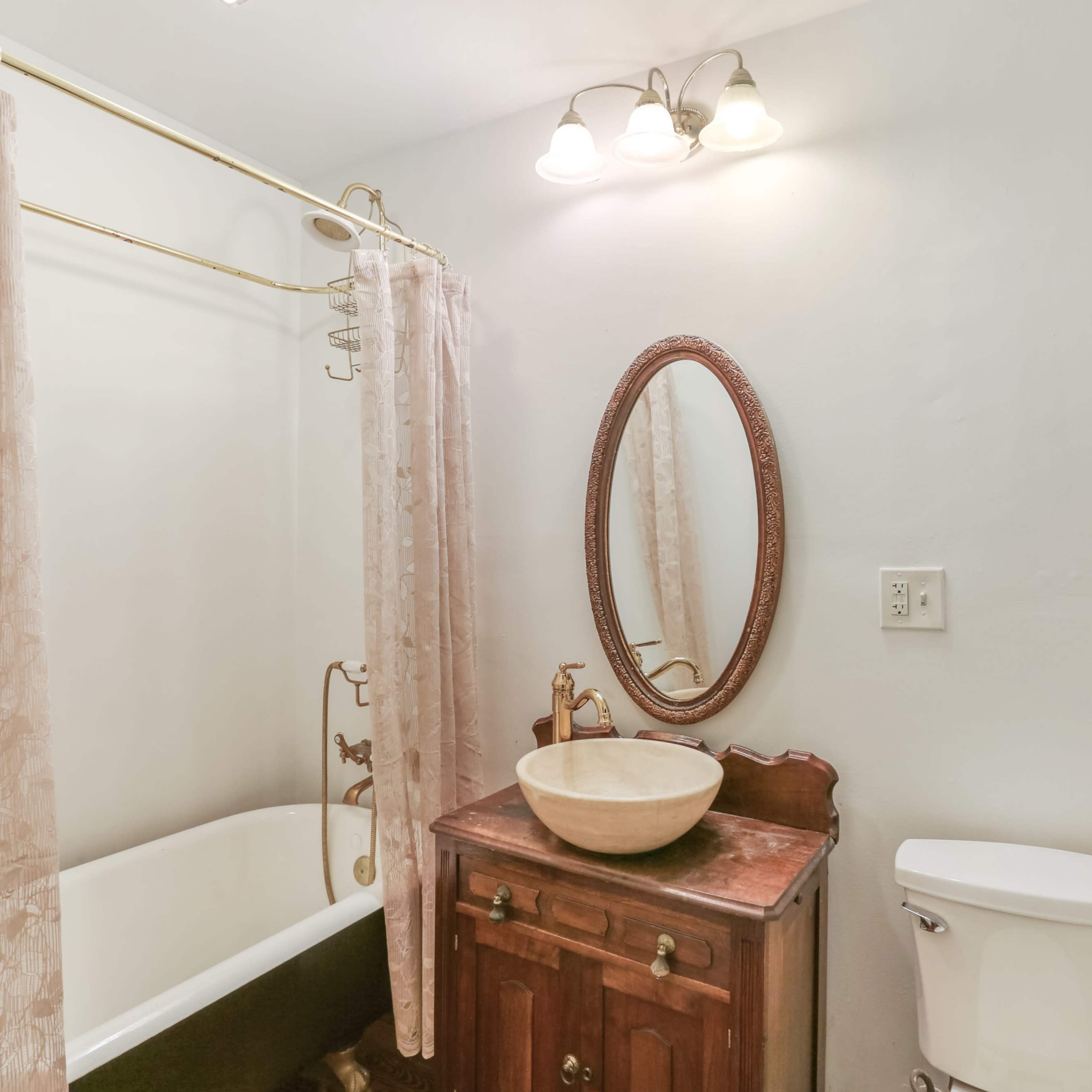 A bathroom featuring a vintage bathtub, a sink on a wooden cabinet, a mirror, and a toilet, all illuminated by overhead lights.