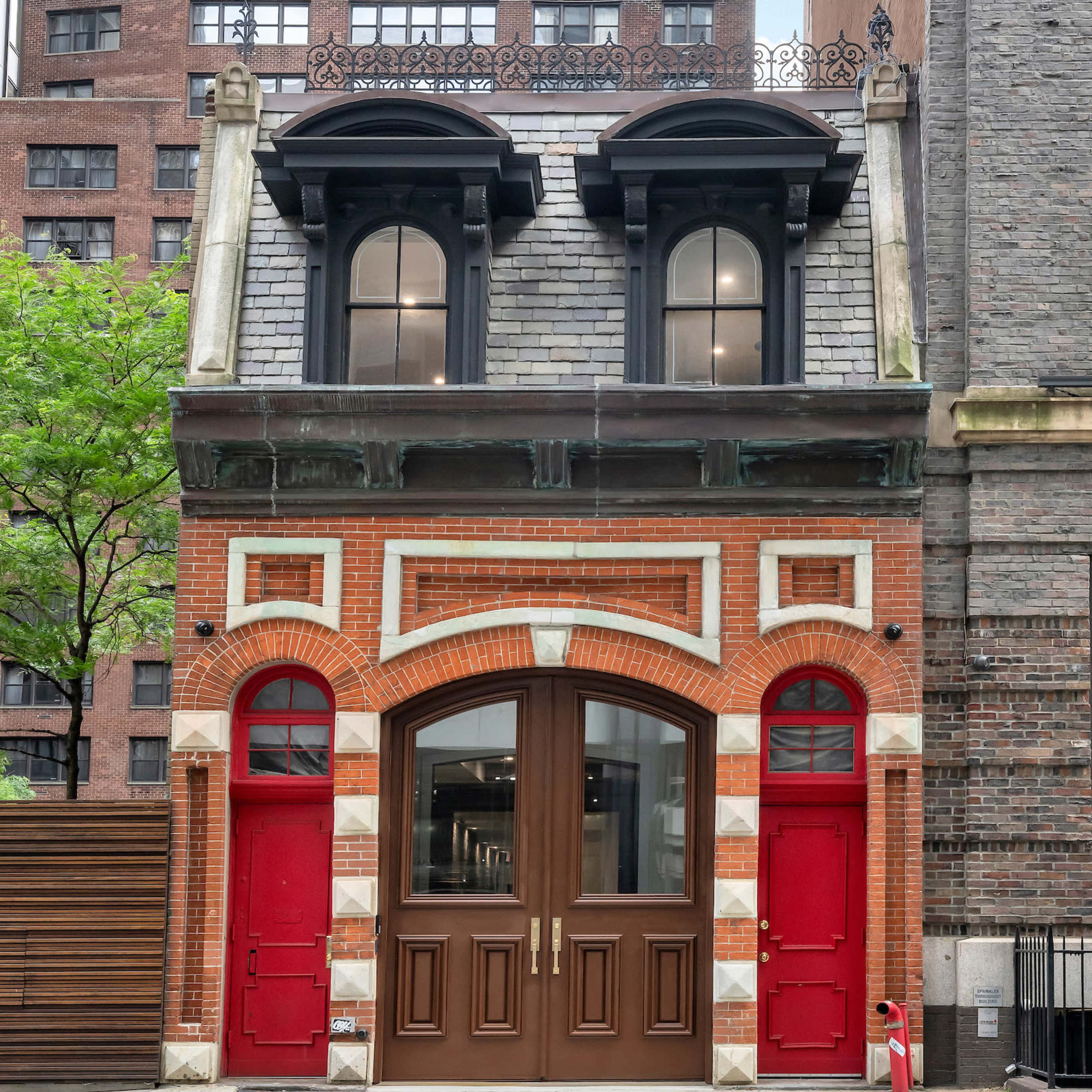 A historic building with a red brick facade, two arched doors, and a decorative roof, situated between modern structures in an urban setting.