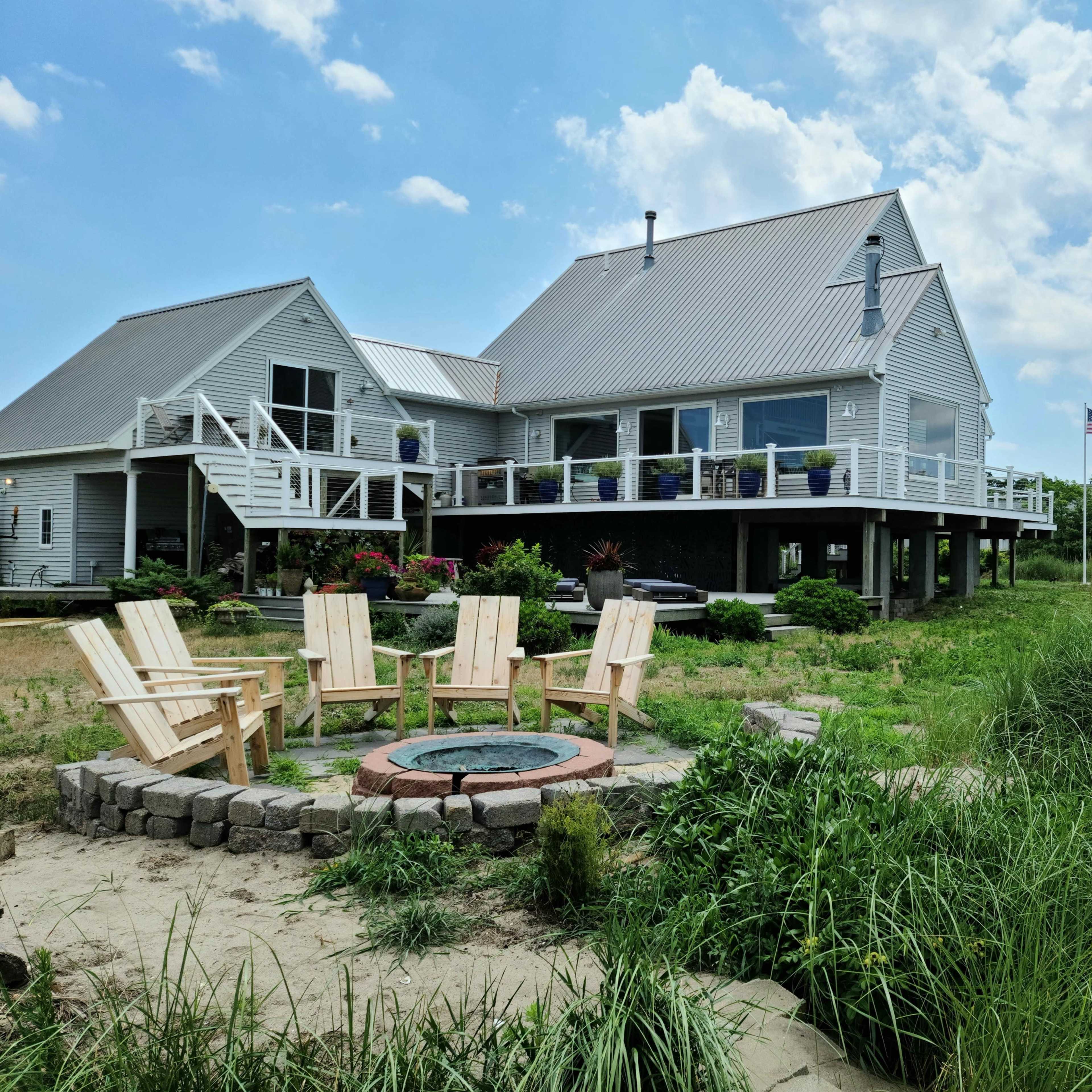 A modern house with a deck overlooks a fire pit surrounded by Adirondack chairs, set against a backdrop of grass and water.
