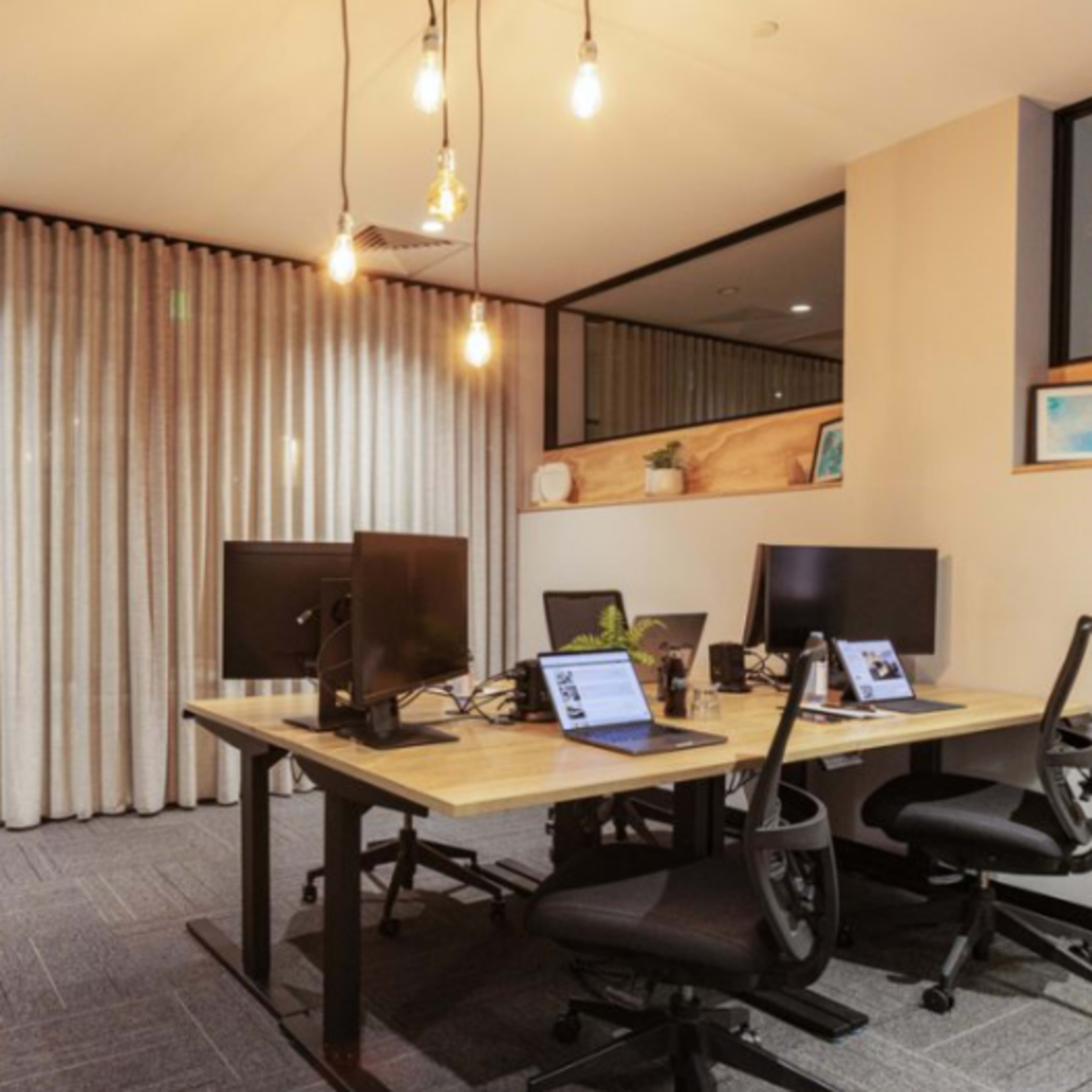 A modern office space featuring several desks with computer monitors, hanging light fixtures, and a wall of curtains.