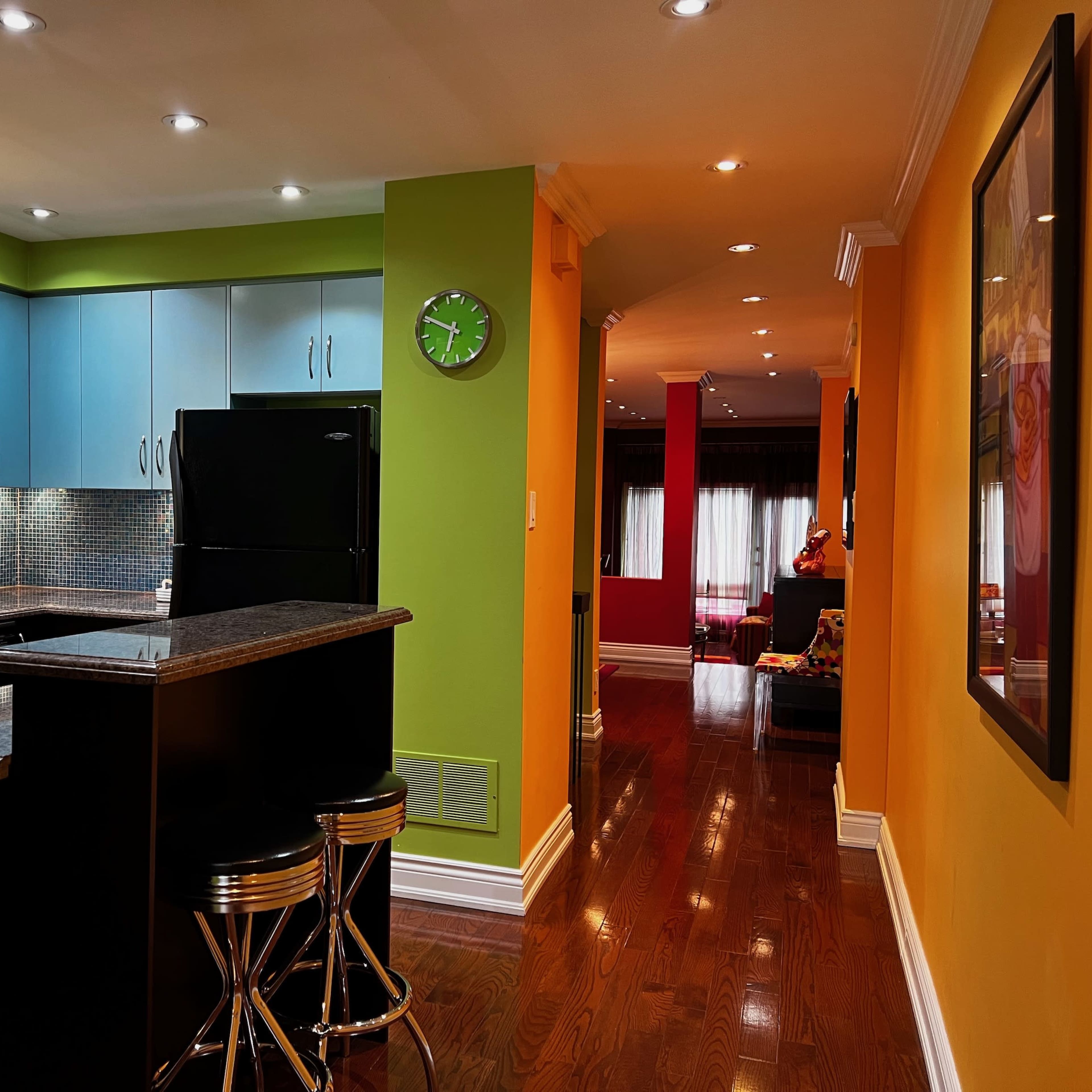 A brightly painted kitchen with blue cabinets and a black refrigerator, leading to a hallway with wooden floors and colorful walls.