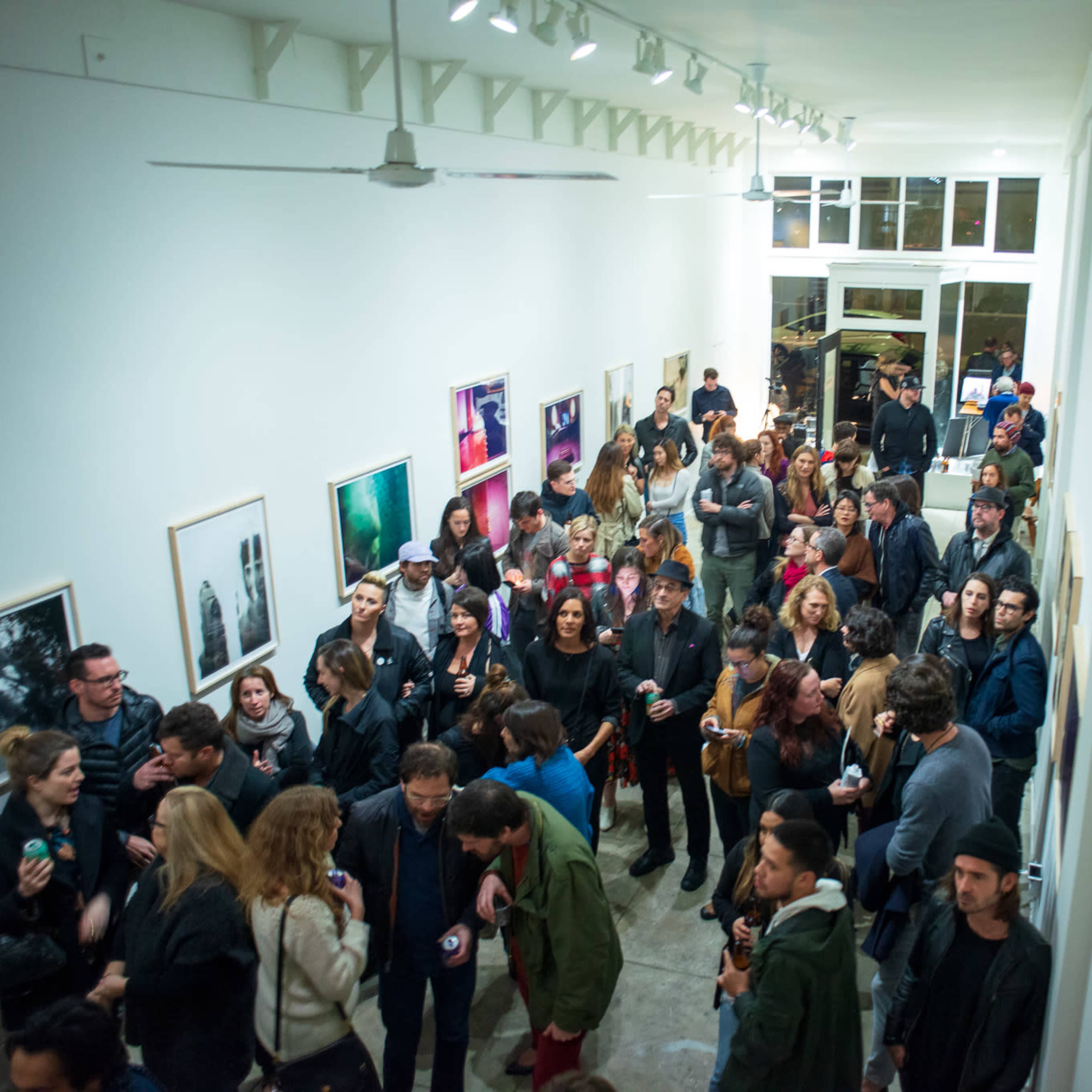 A large crowd gathers in an art gallery, surrounded by framed photographs displayed on the white walls.