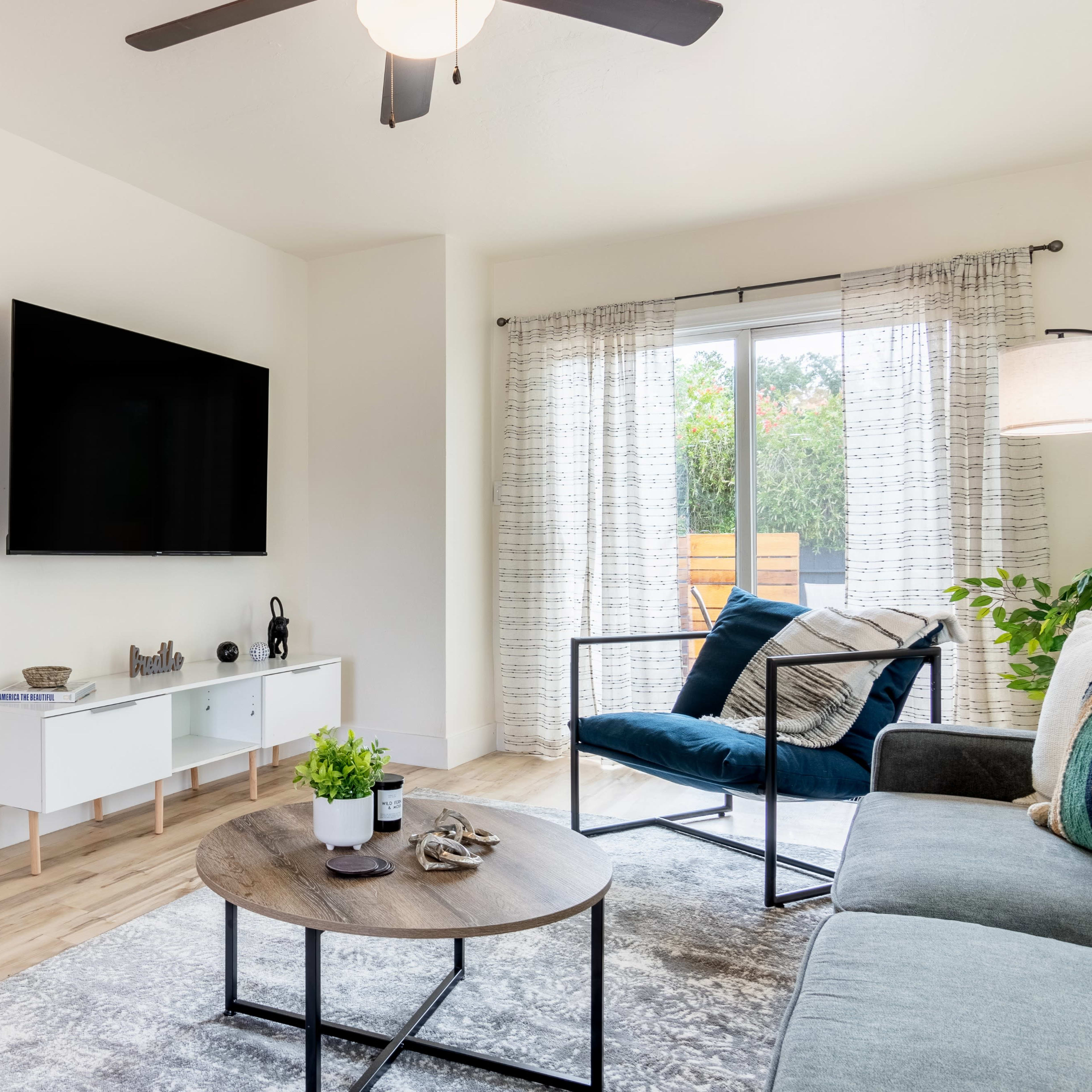 The living room features a television mounted on a white wall, a round coffee table, and a blue sofa, with natural light streaming in through large sliding glass doors.
