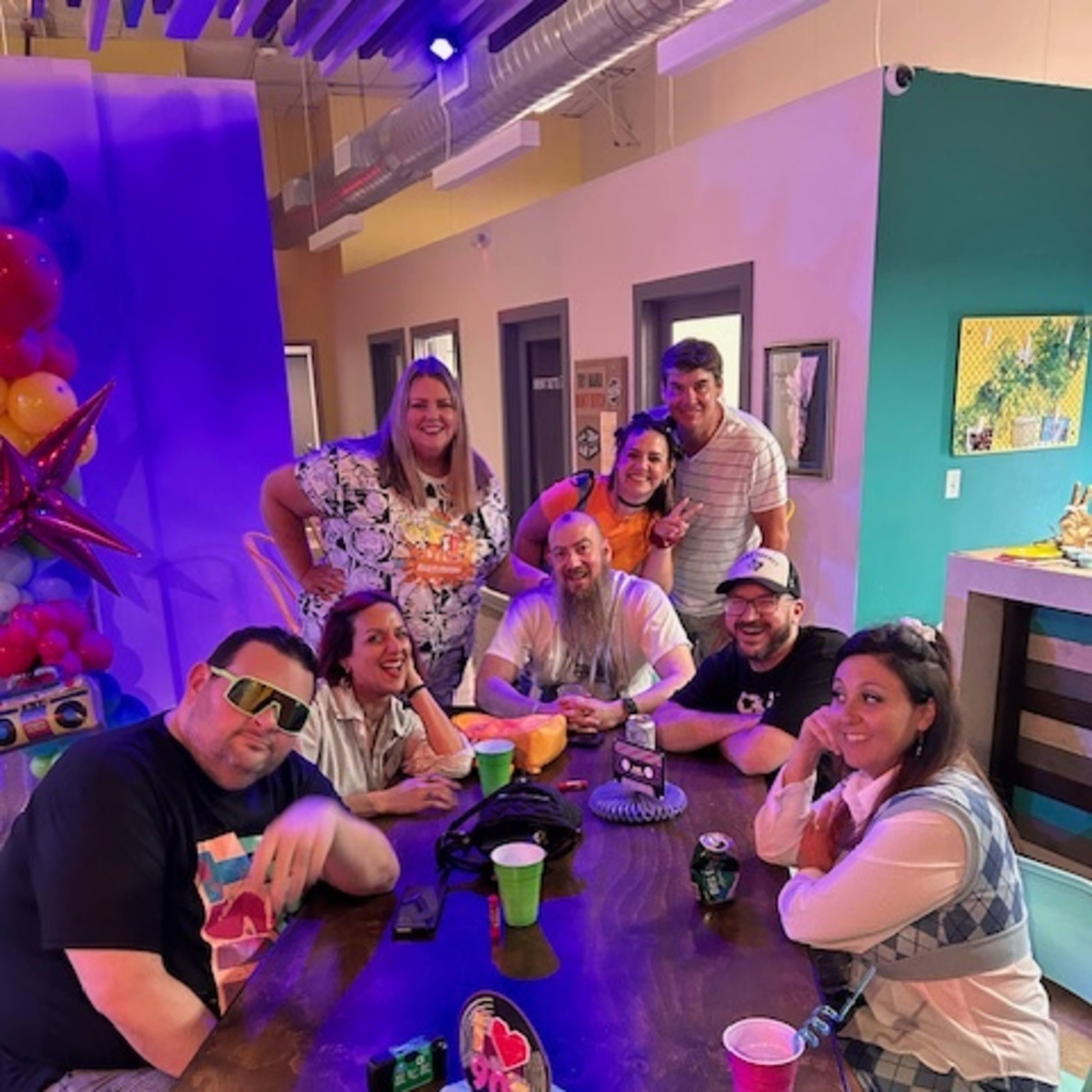 A group of eight people poses around a table, smiling and holding drinks in a colorful indoor setting.