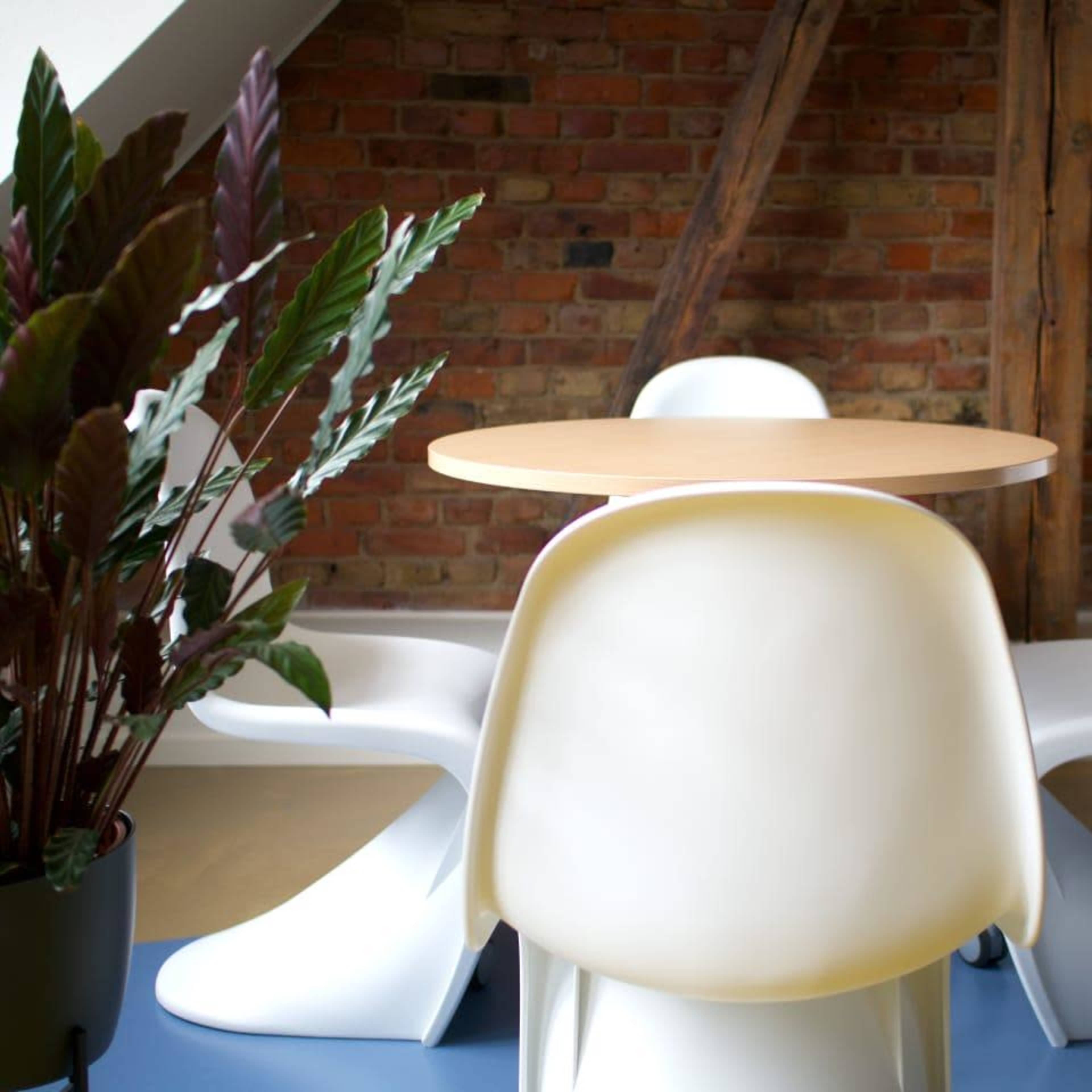 A round wooden table is set in a room with exposed brick walls, accompanied by three white chairs and a potted plant in the foreground.