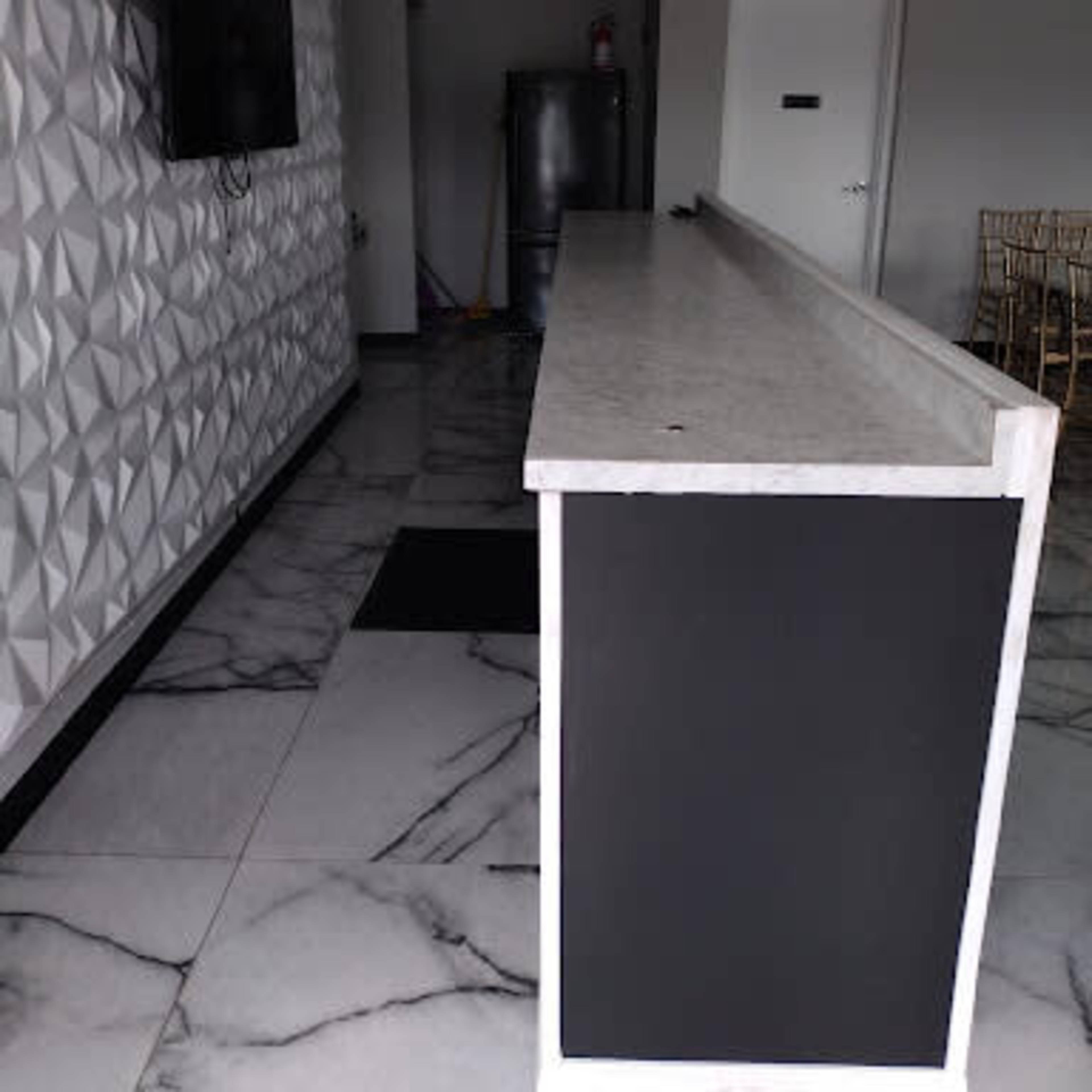 A bar with a marble countertop along a wall featuring textured geometric patterns, set in a room with tiled flooring and a wall-mounted television.