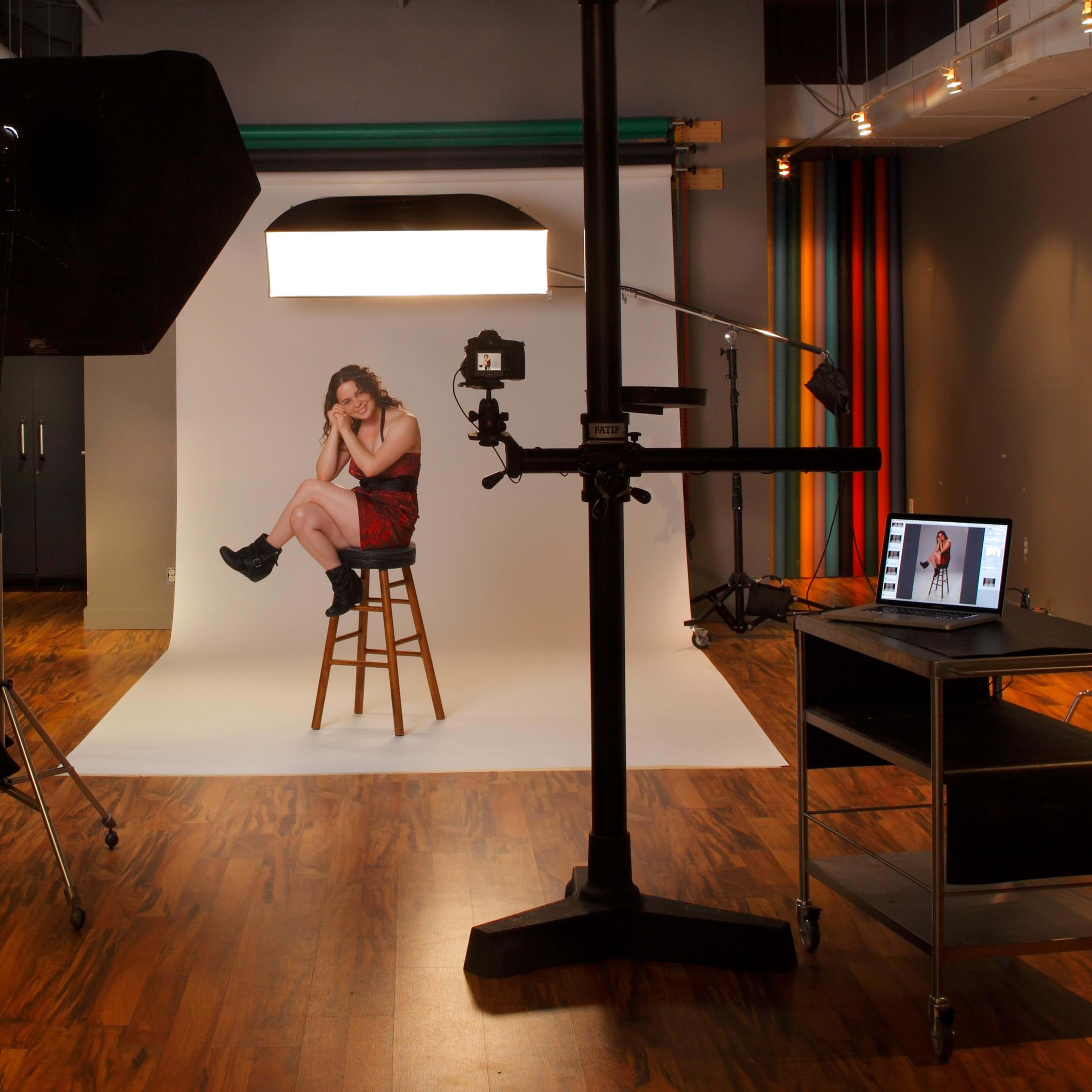 A model sits on a stool in a photography studio, surrounded by lighting equipment and a computer displaying an image.