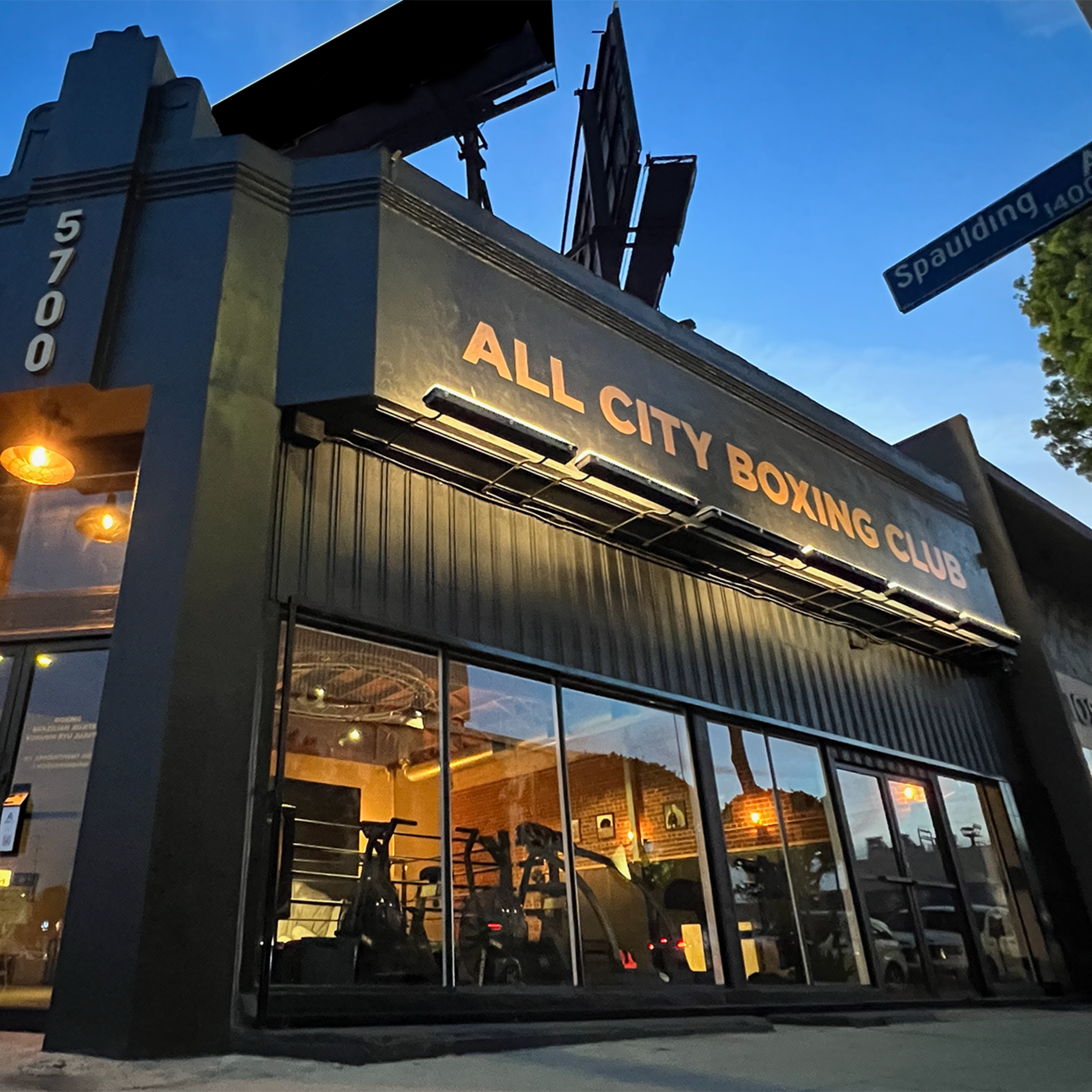 The exterior of the All City Boxing Club, featuring large windows and a sign, set against a twilight sky.