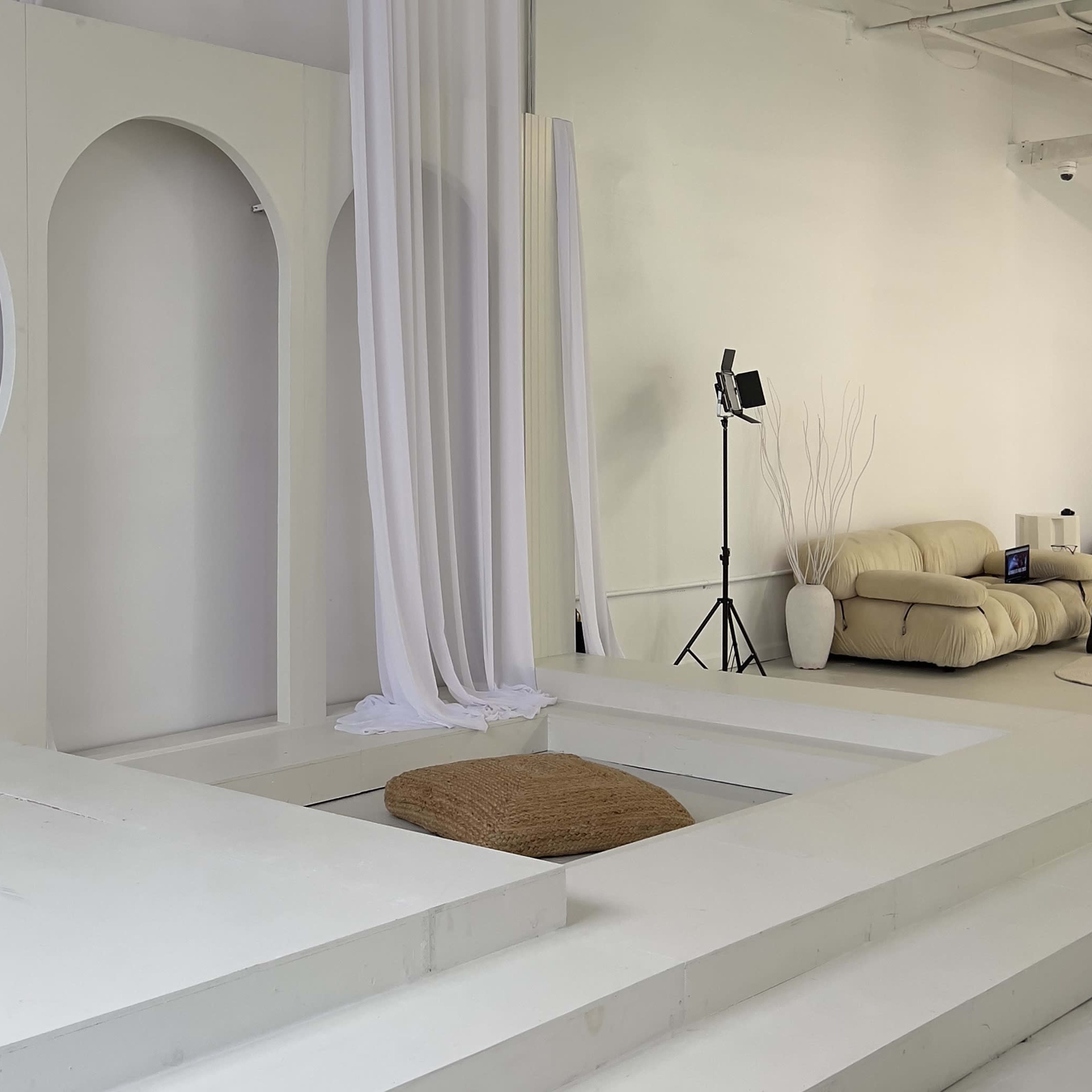 A minimalist interior space with white walls, a circular window, steps leading to a sunken area with a circular mat, and a beige sofa in the corner.