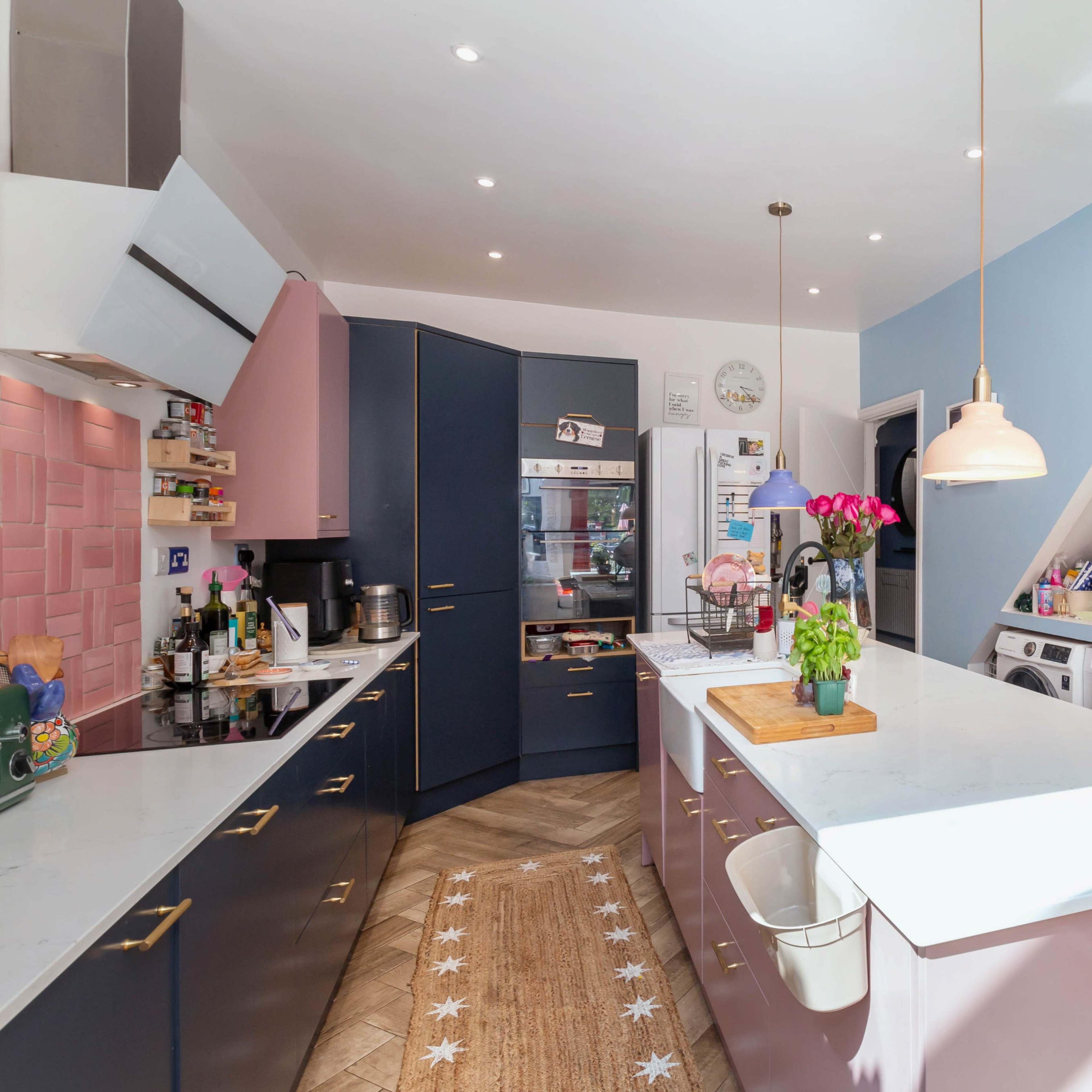 A modern kitchen features a combination of navy blue and pink cabinets, a central island with a cutting board, and a wall with a light-up sign that reads "LET'S STAY HOME."