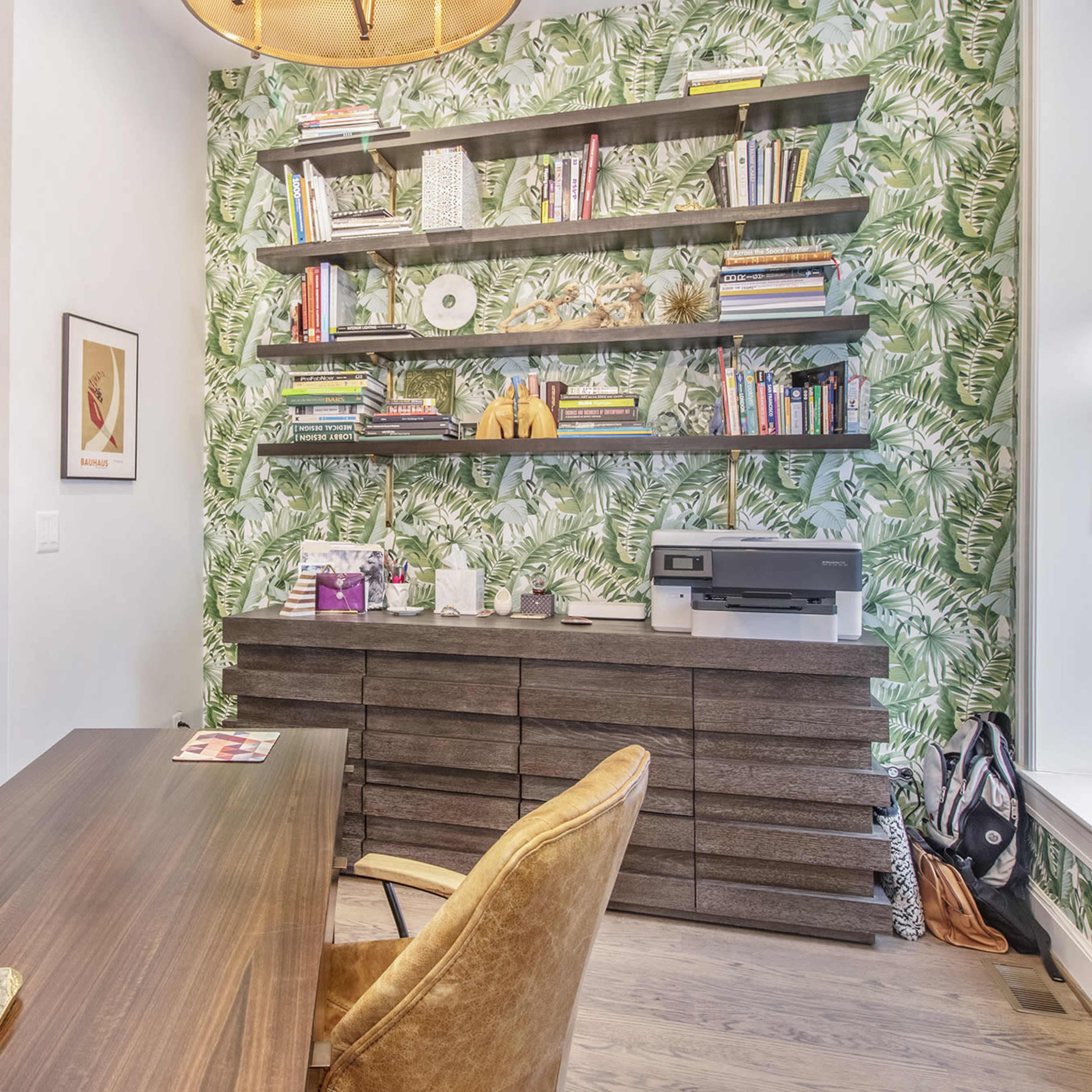 A home office features a wooden desk and chair, green leaf-patterned wallpaper, and shelves filled with books and decorative items.