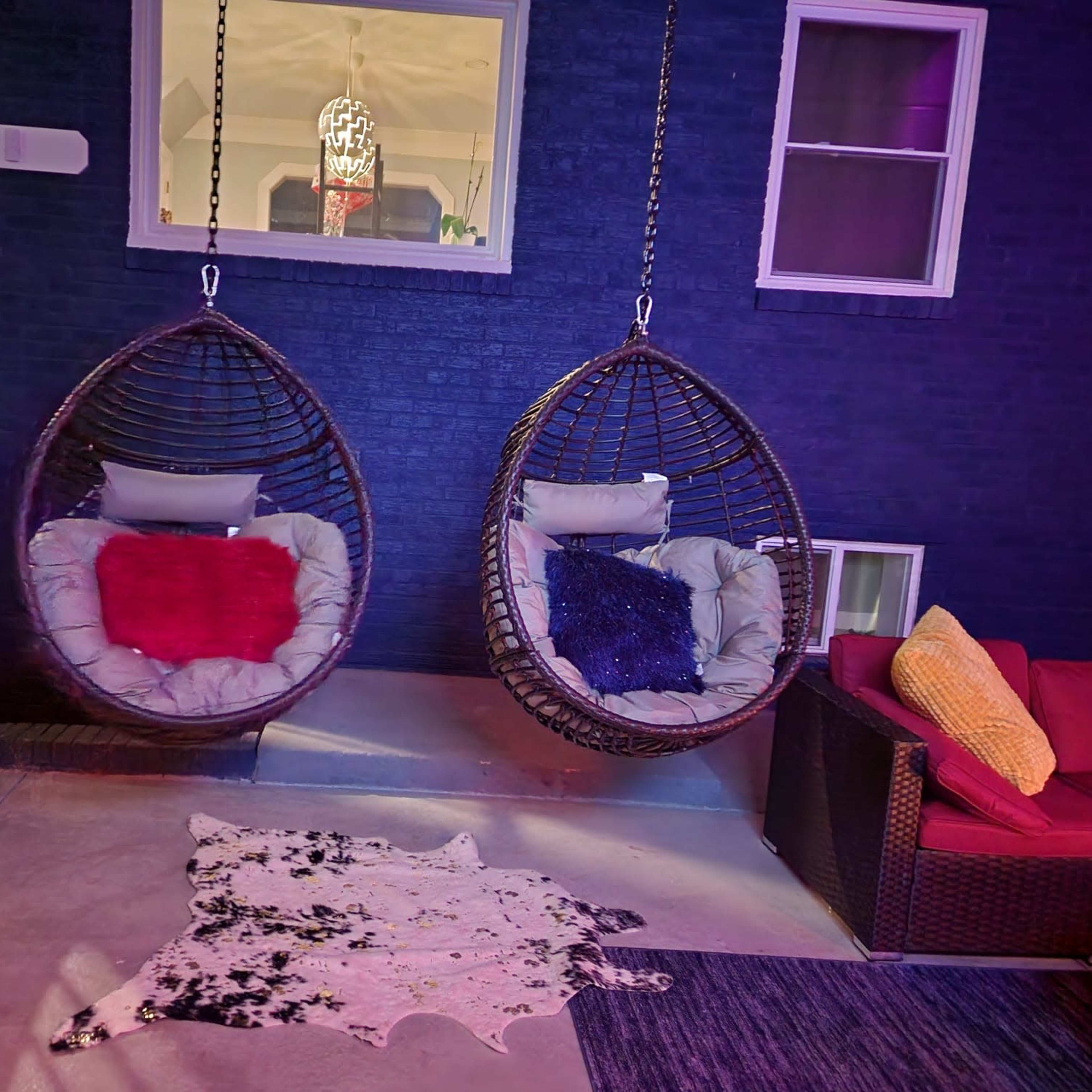 Two hanging chairs with cushions attached to a wall in a cozy outdoor space, accompanied by a decorative rug and a red couch.