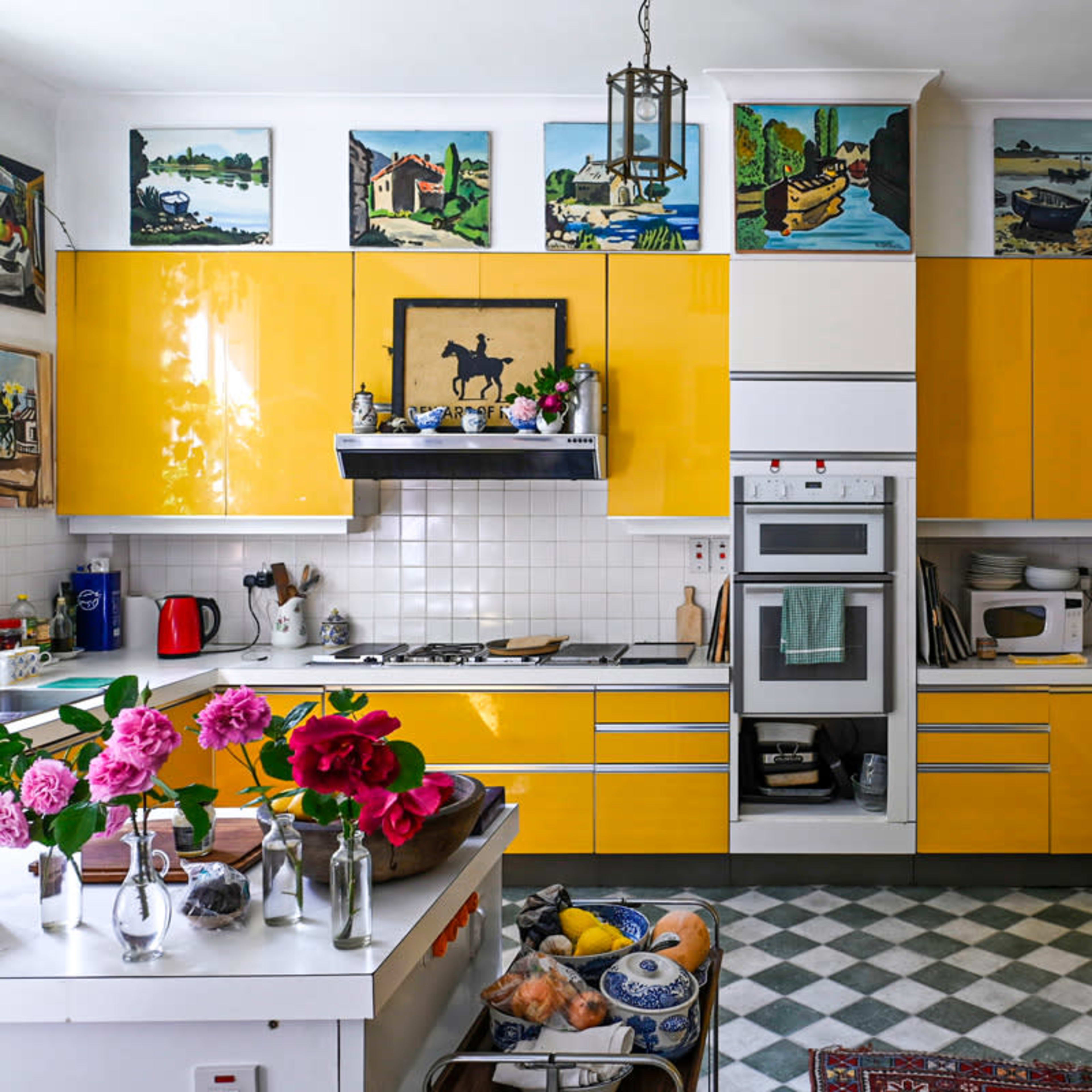 The kitchen features bright yellow cabinets, a checkerboard floor, and various paintings on the walls, with flowers and fruit displayed on the countertops.