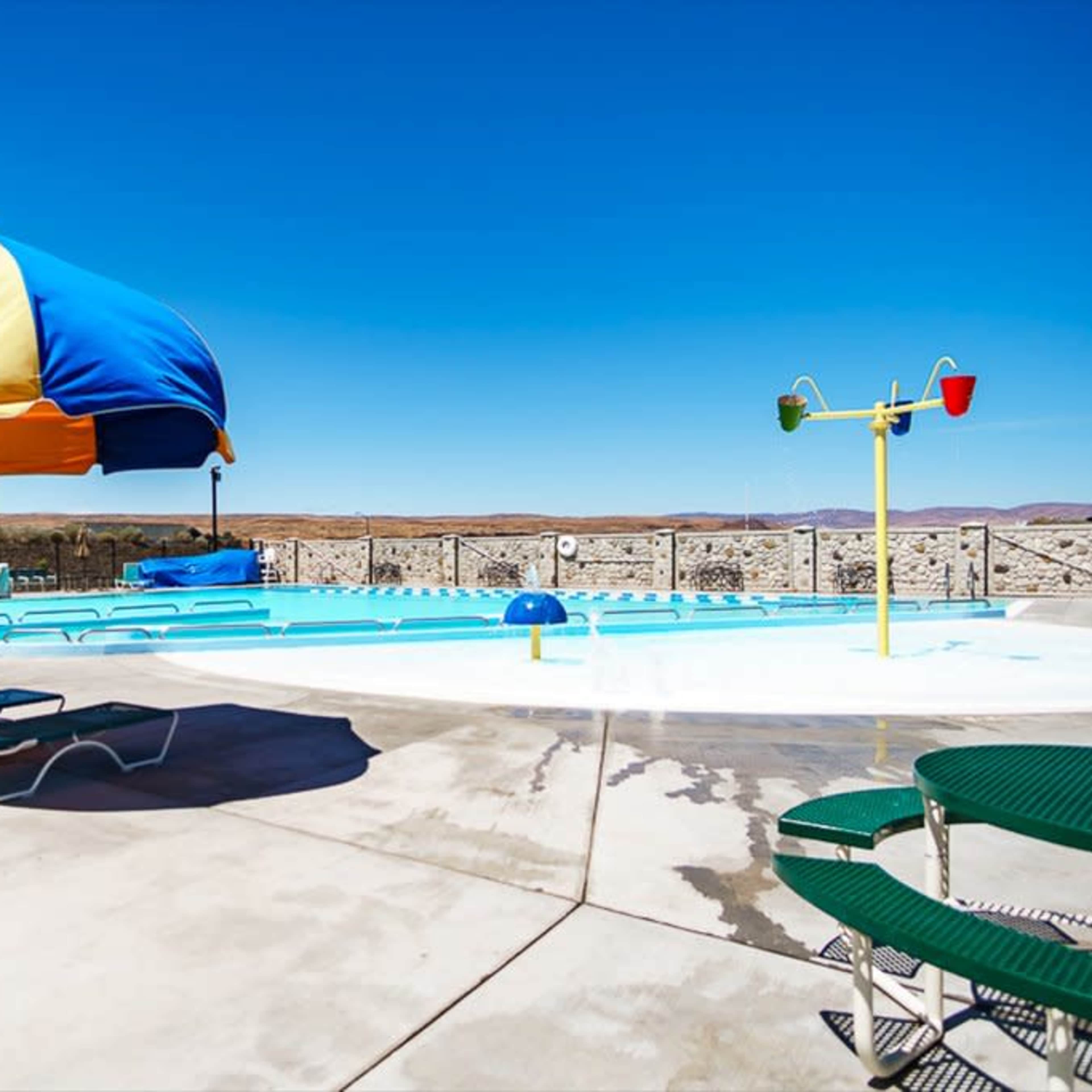 A bright pool area features a blue and yellow umbrella, green lounge chairs, and a shallow swimming pool under a clear blue sky.