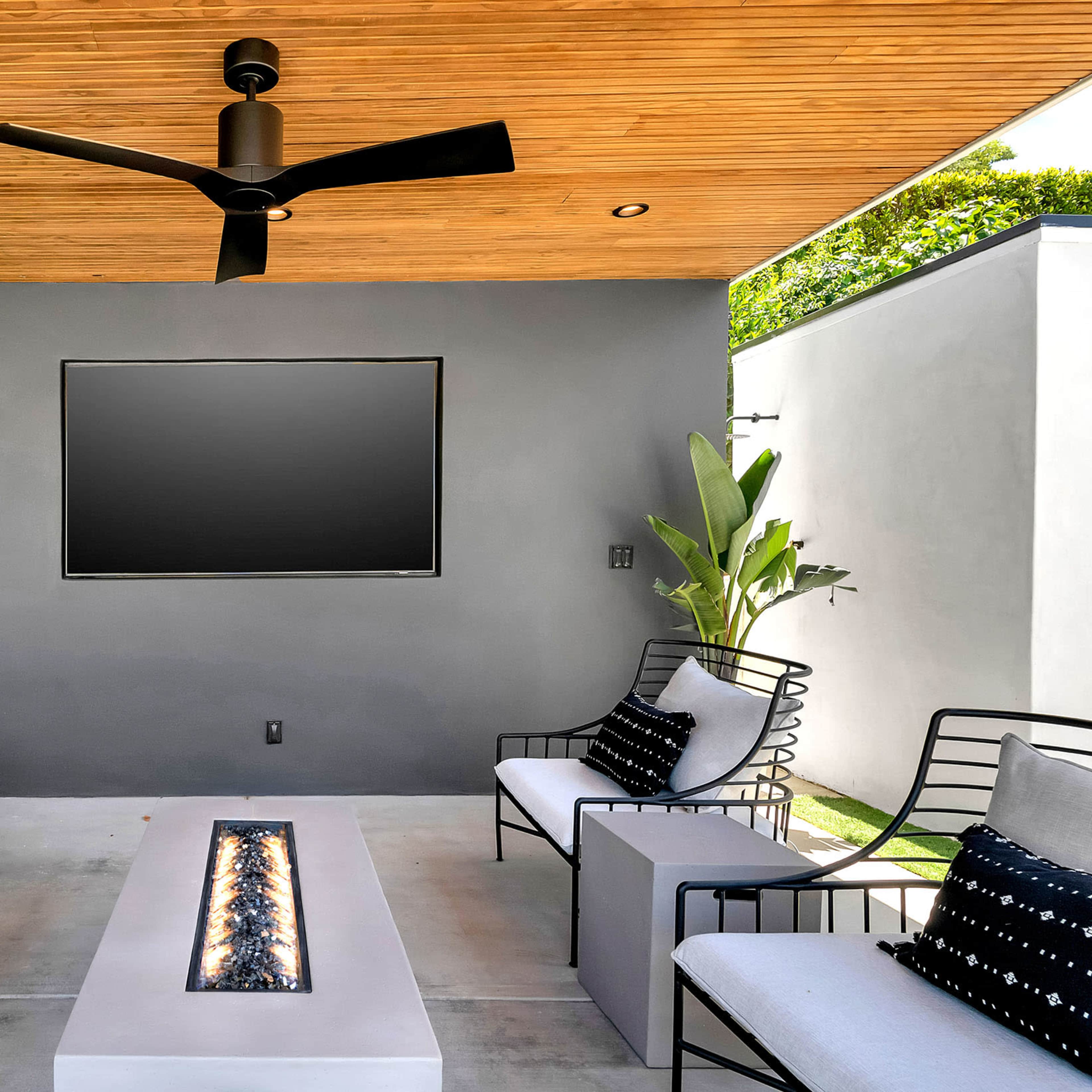 A modern outdoor lounge area with two seating benches, a central fire feature, a television on the wall, and greenery in the background.