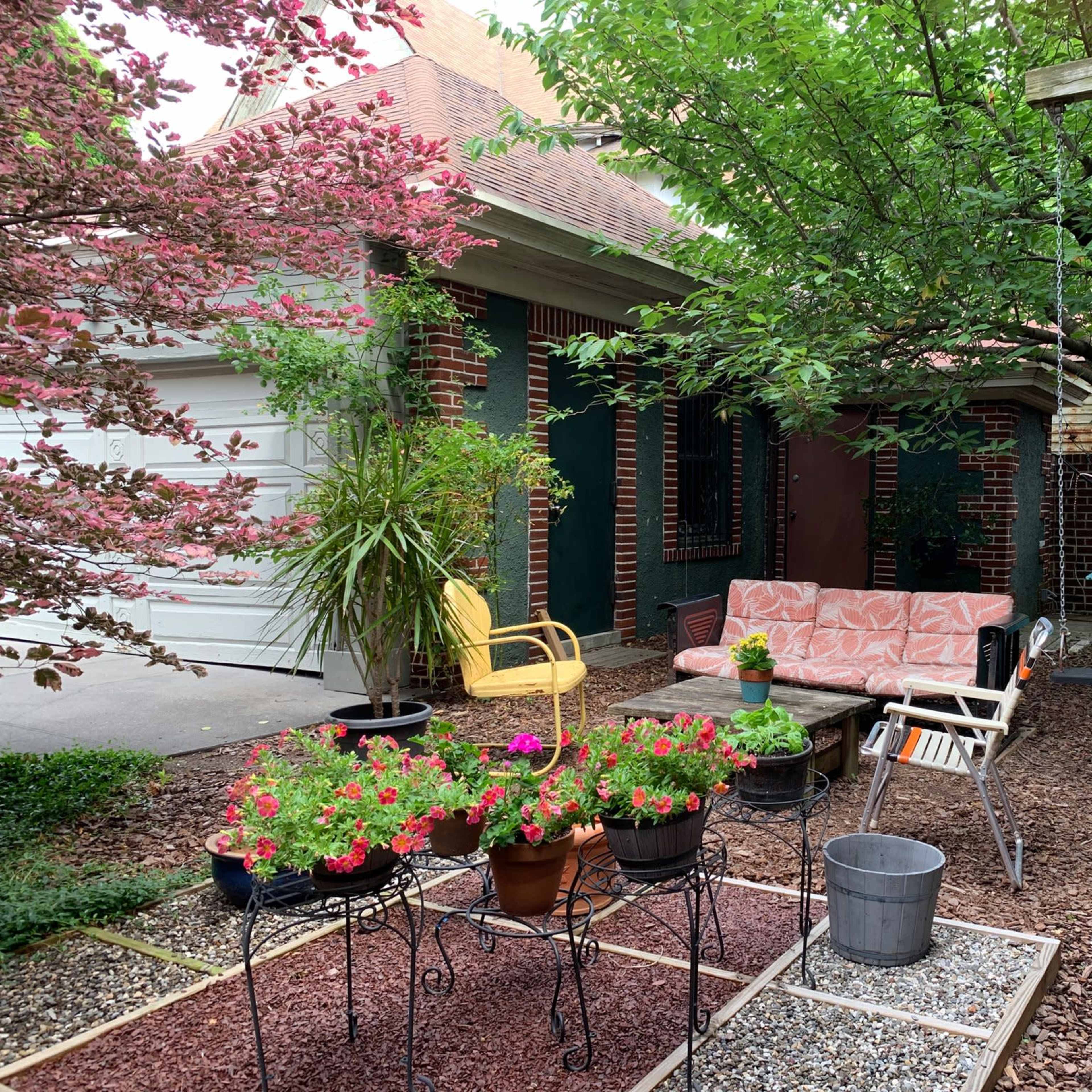 A cozy outdoor garden area features a seating arrangement, potted flowers, and a gravel path surrounded by greenery.