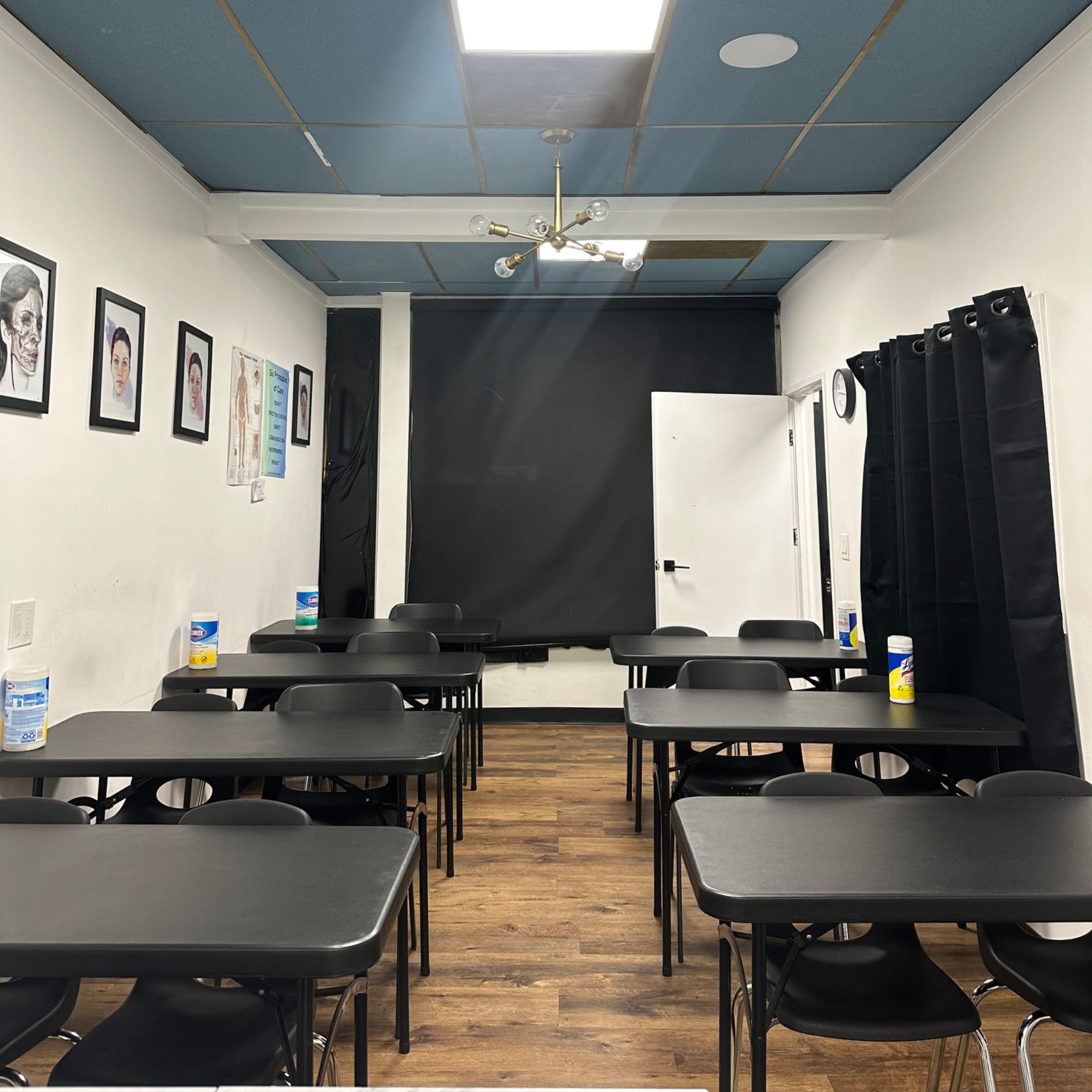 A classroom setup with multiple black tables and chairs, a projector mounted on the ceiling, and black curtains covering a window.