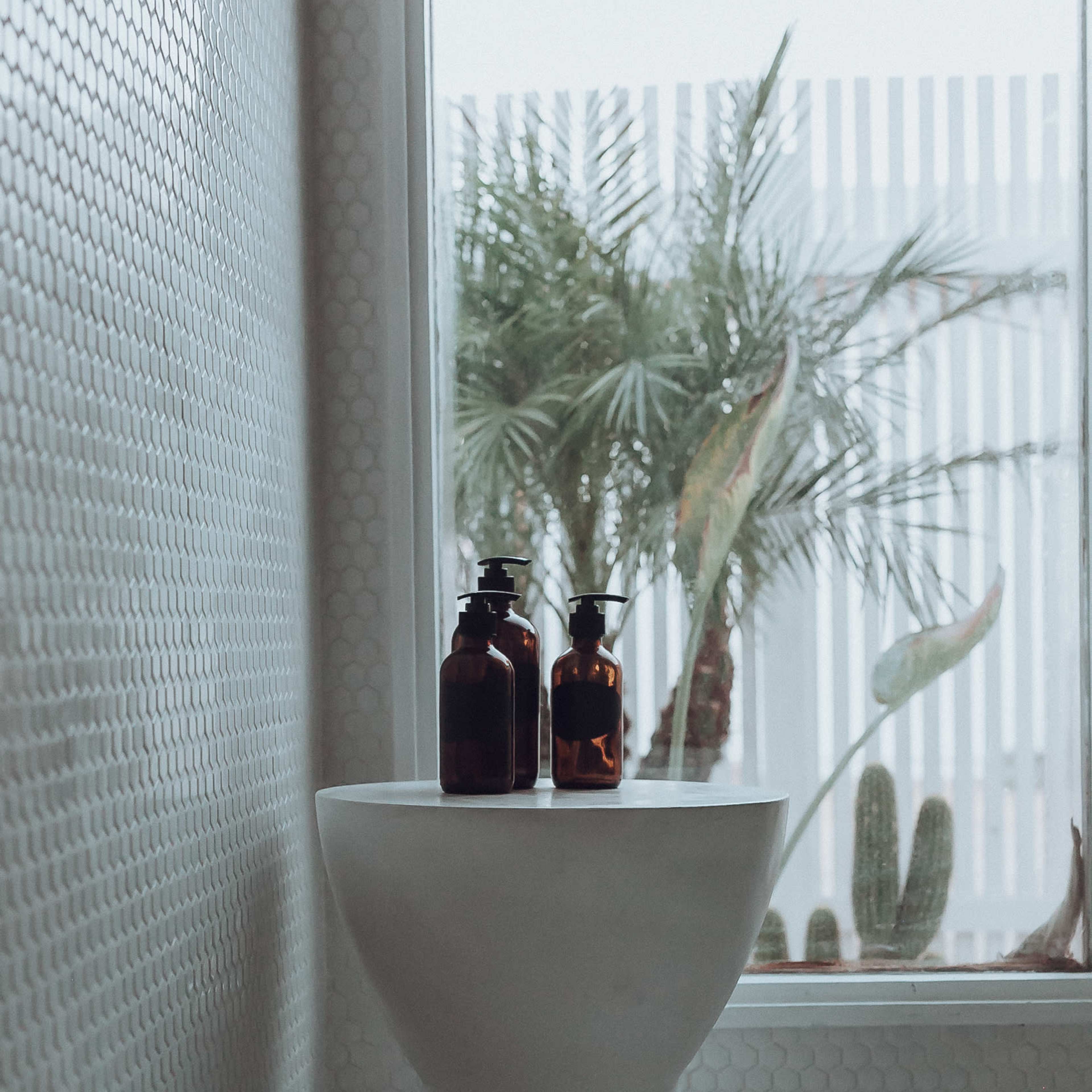 A modern bathroom features a light-colored, sculptural stool holding two brown glass bottles, with a palm plant visible through a window.