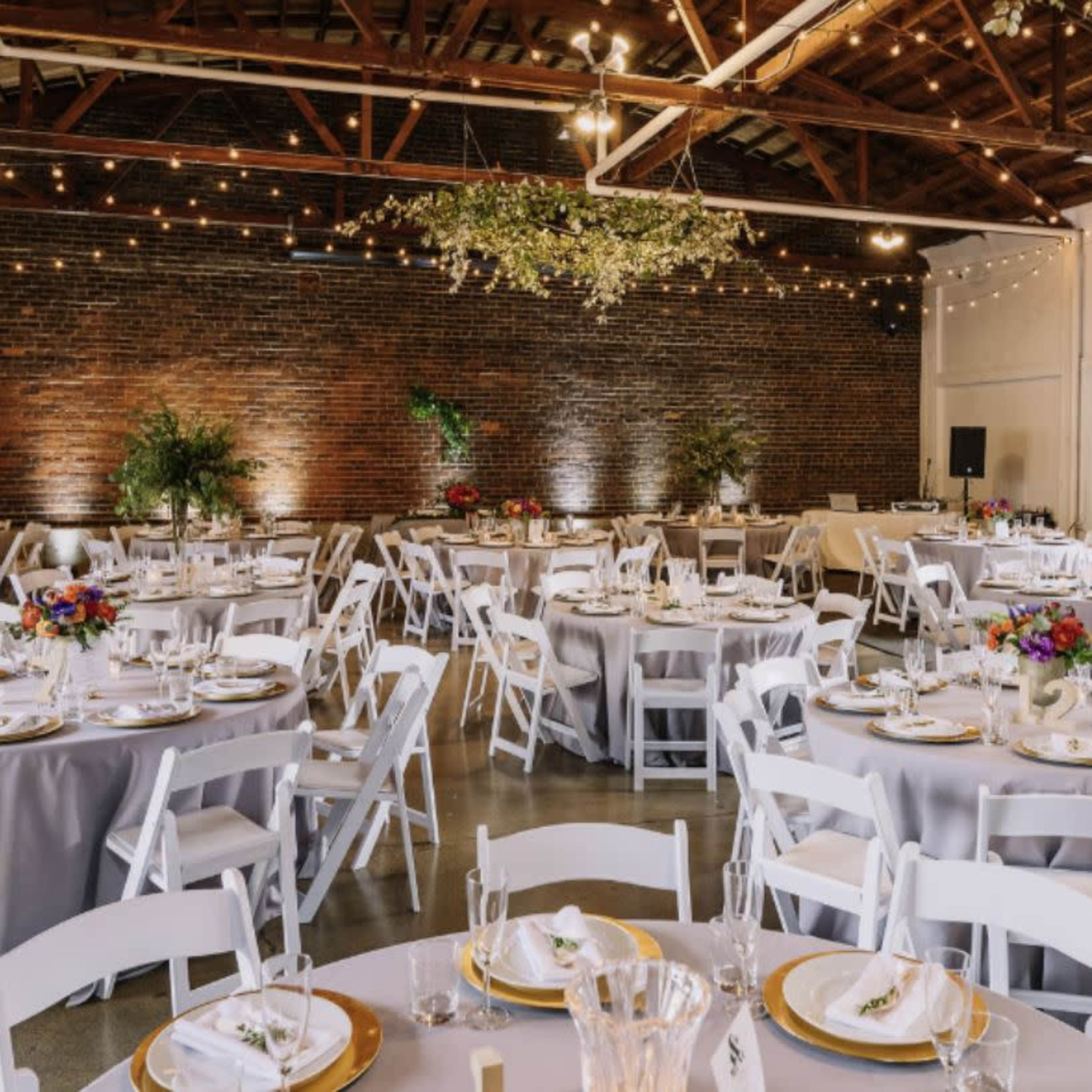 A spacious event venue features round tables set with white chairs and gray table linens, adorned with floral centerpieces and overhead string lights.
