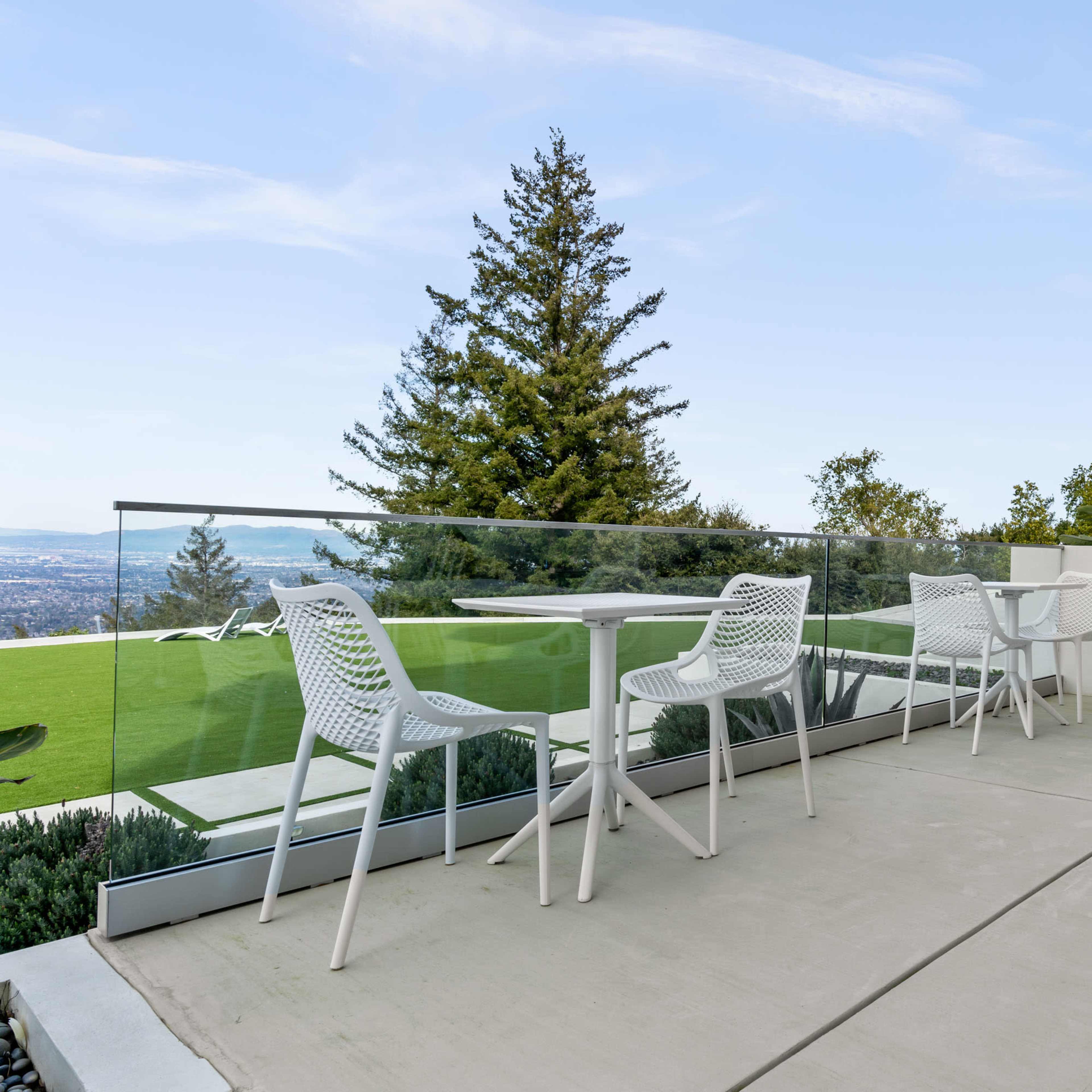 A modern terrace features white chairs and a table with a clear glass railing overlooking a green landscape and distant hills.