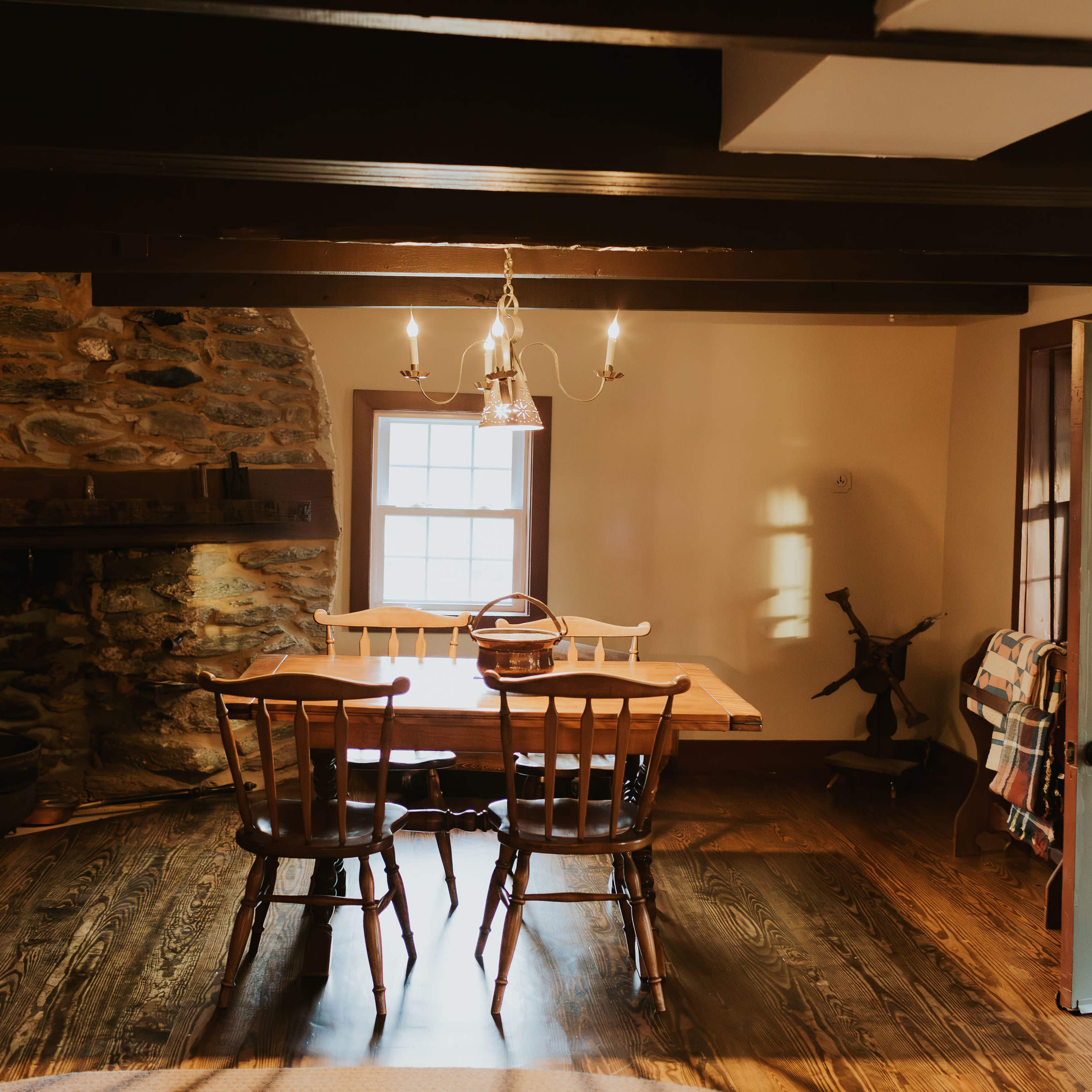 A rustic dining area with a wooden table, four chairs, and a stone wall, illuminated by natural light coming through a window.