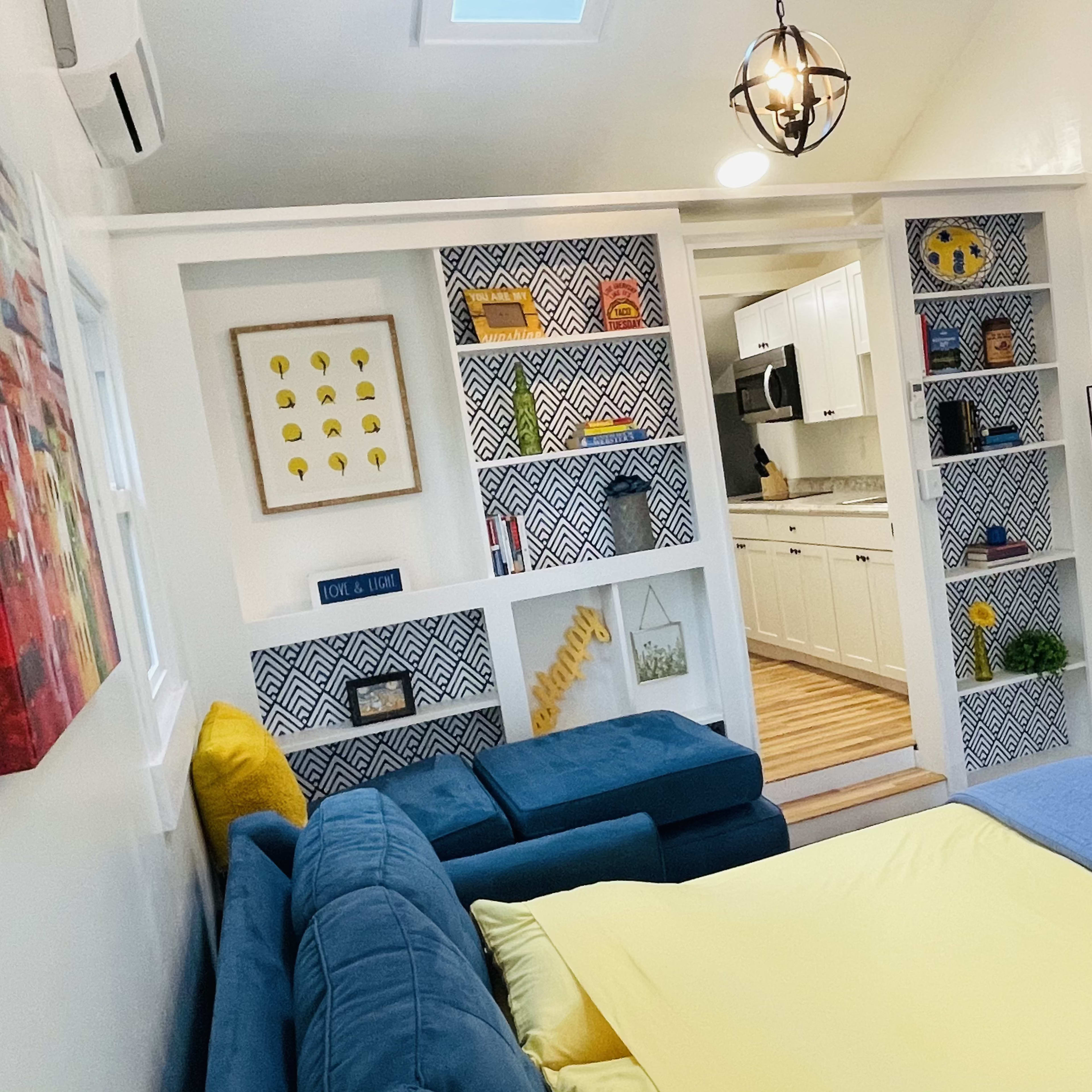A living area with a blue couch, yellow bedding on a pull-out bed, and built-in shelves displaying decorative items and books, leading into a kitchen space.