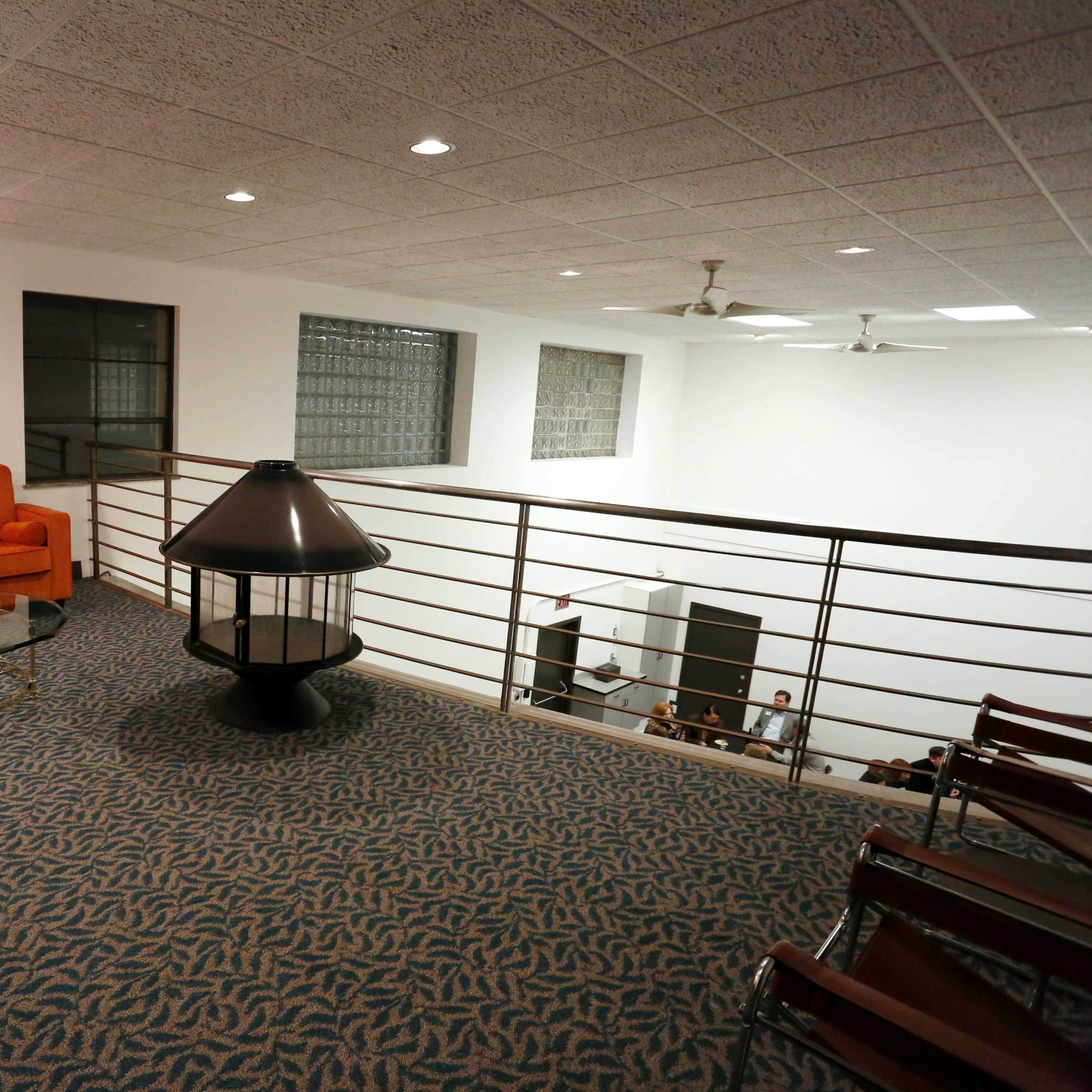 A spacious, well-lit mezzanine features an orange couch, a glass coffee table, and a lamp, overlooking a room with chairs and people.