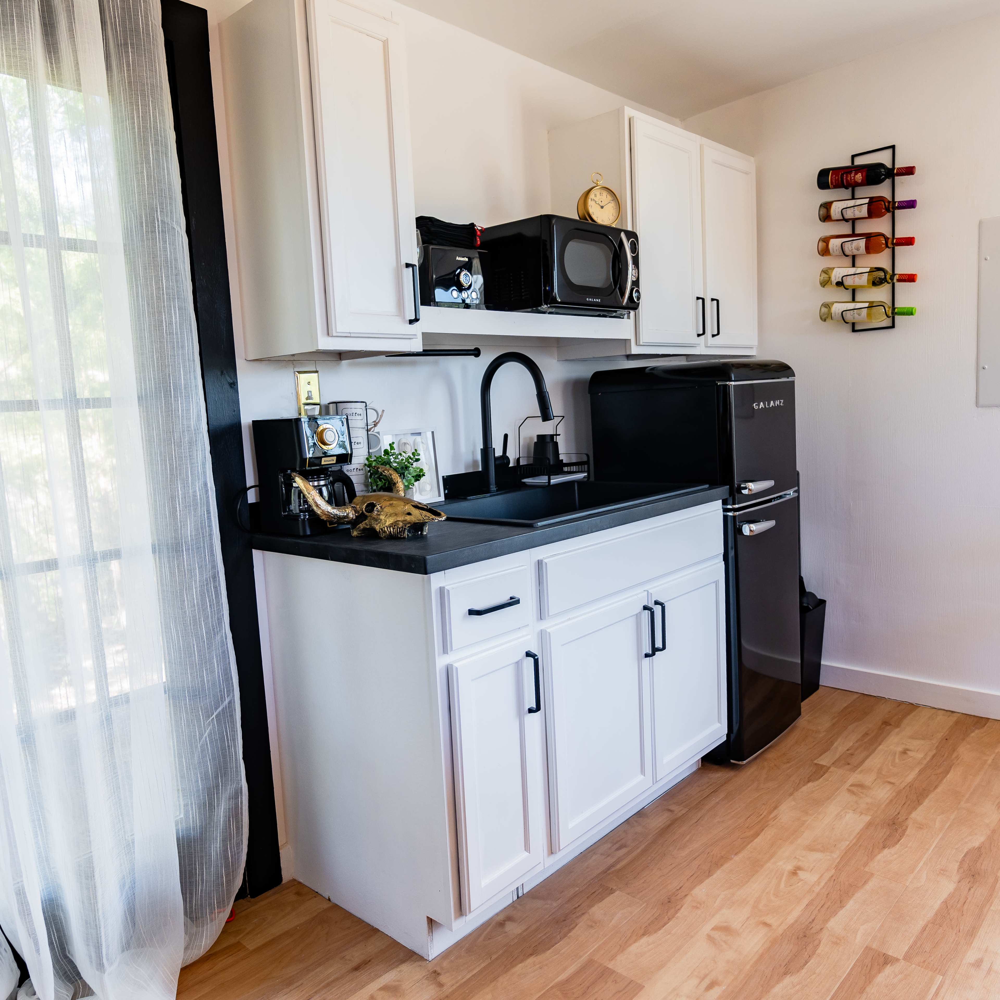 A small kitchen features white cabinets, a black countertop, a microwave, and a compact refrigerator next to a window draped with sheer curtains.