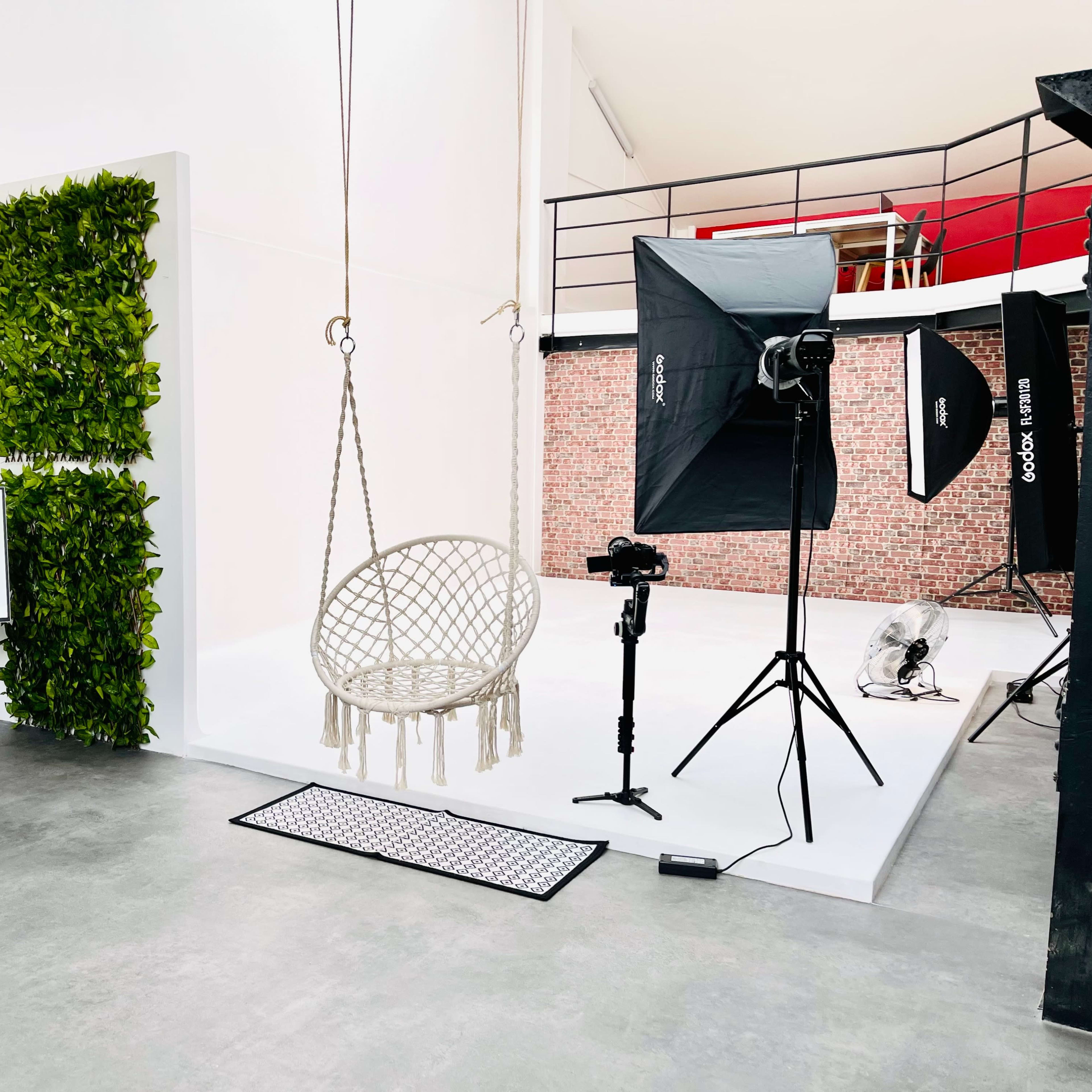A minimalistic studio space featuring a hanging chair, a backdrop of greenery, and equipment for photography, including lights and a camera on a tripod.