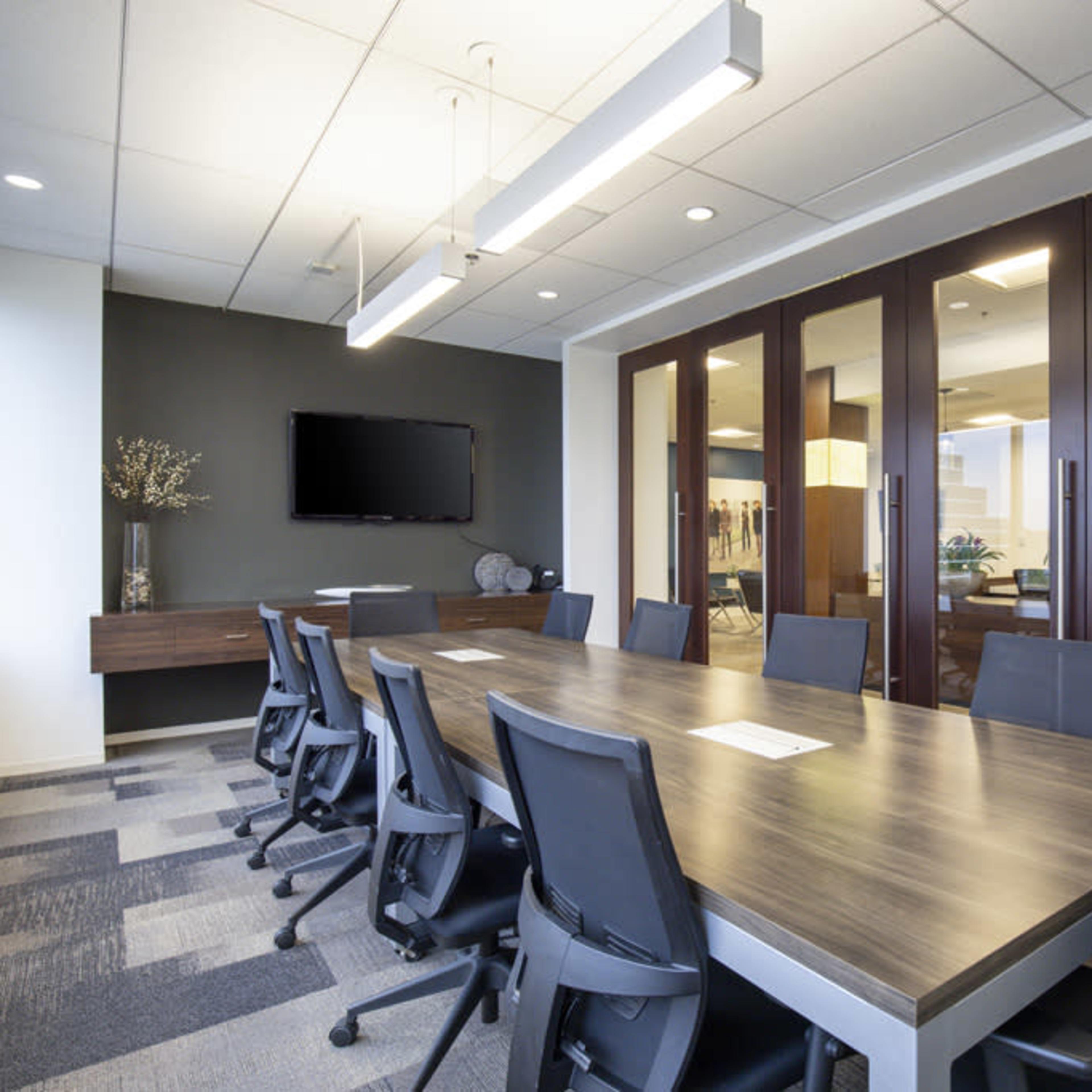 A modern conference room features a long wooden table surrounded by ergonomic chairs, with large glass doors leading to another office space.