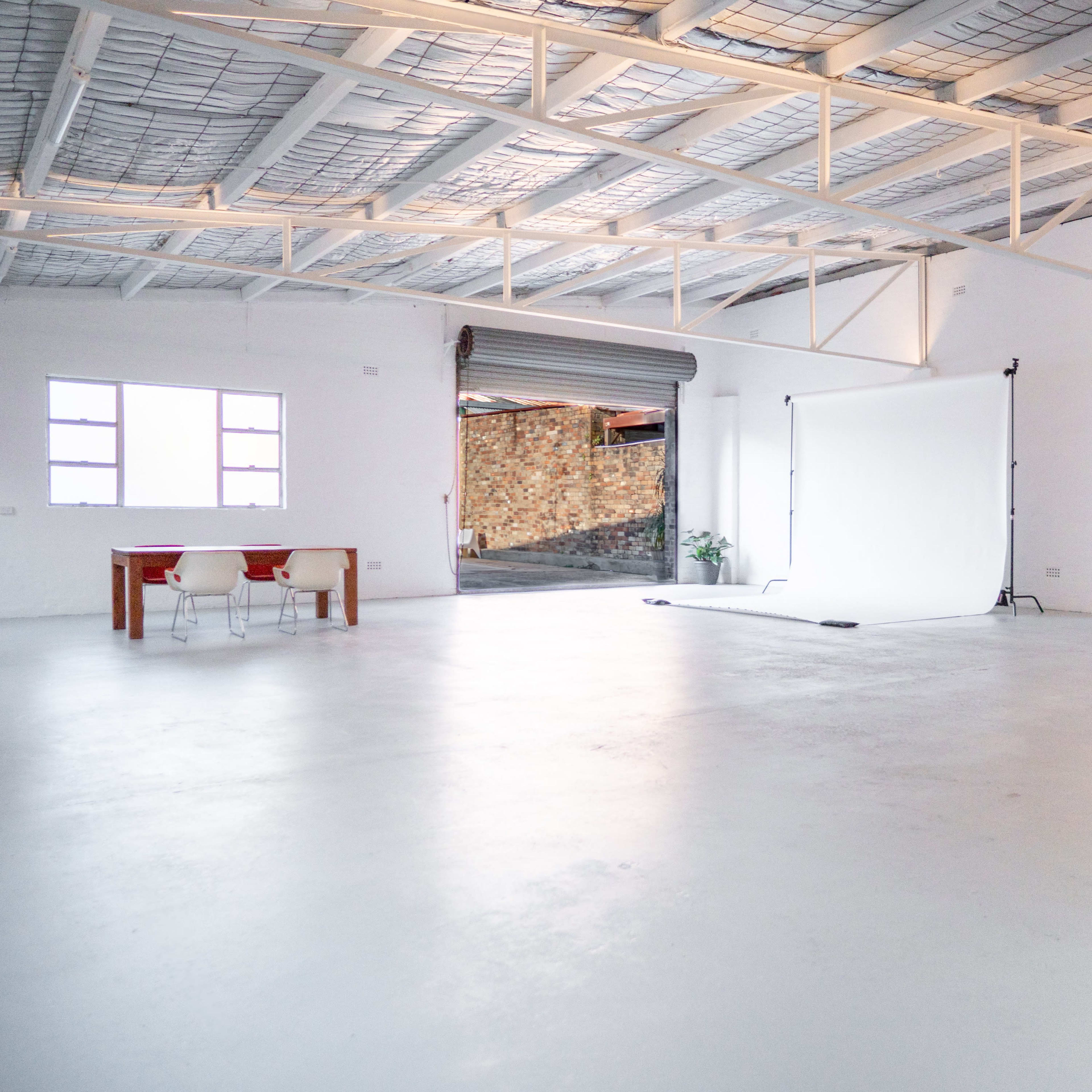 A spacious, empty industrial-style room with a white backdrop set up, a table, and two chairs.